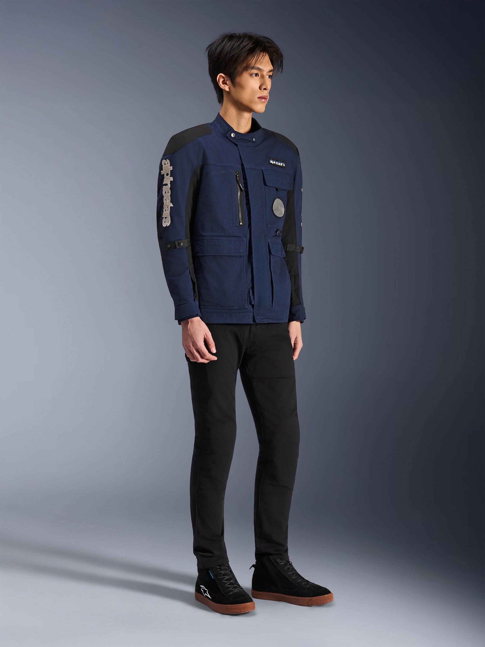 Alpinestars Dice Jacket, donkerblauw en zwart, moderne motorjas in veldstijl gedragen door een model met zwarte broek en zwarte hoge sneakers, met meerdere utility-zakken, verticale Alpinestars-branding op de mouw en geweven patches, urban eigentijdse motorkleding