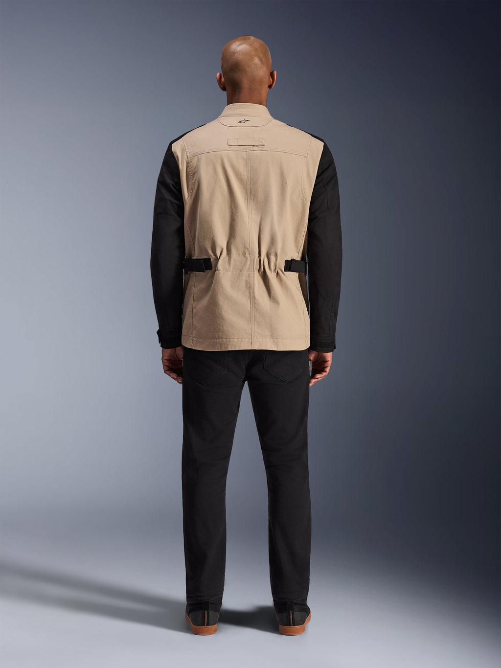 Alpinestars Dice Jacket, urban motorjas, tan en zwart, achteraanzicht op model, voorzien van polykatoen twill-constructie, verstelbare taillebanden en een klein geborduurd logo op de kraag, gedragen met een zwarte broek