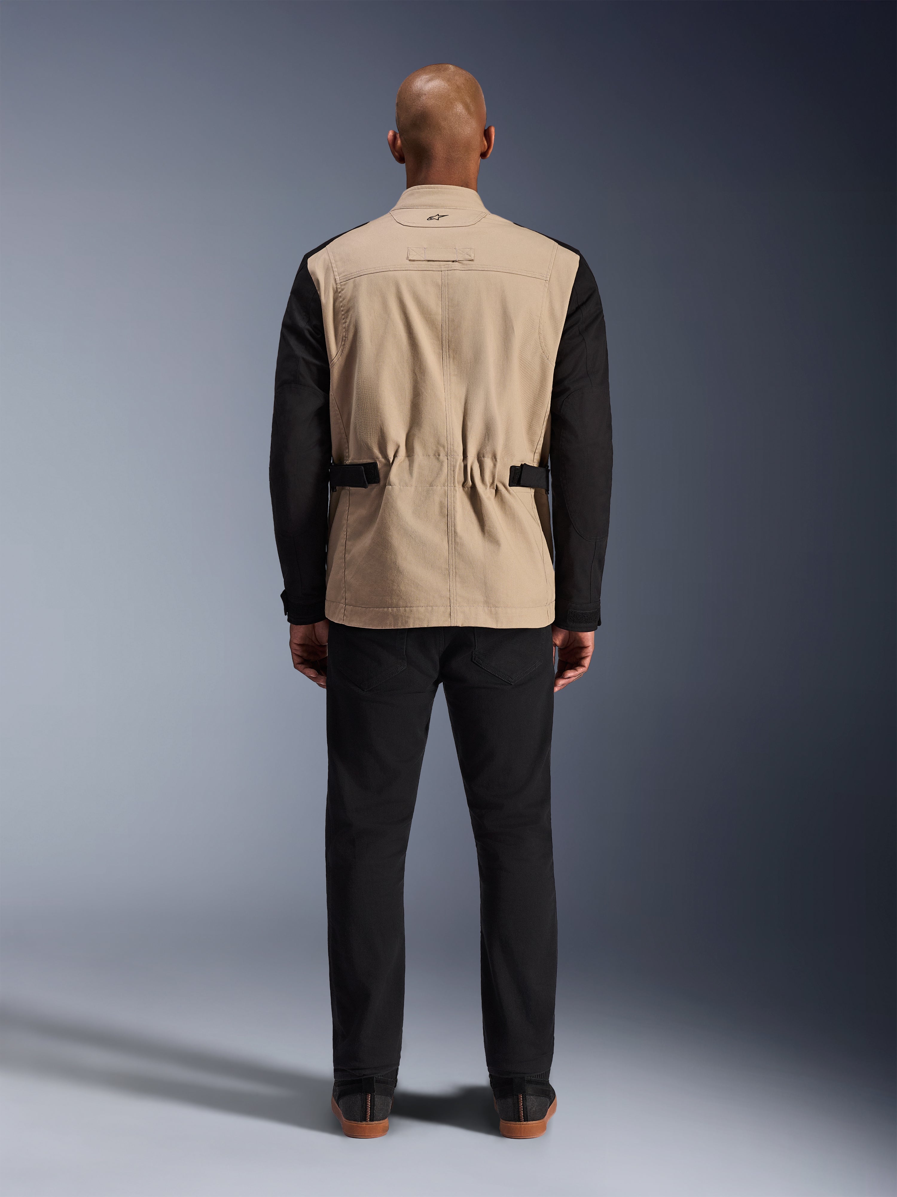 Alpinestars Dice Jacket, urban motorjas, tan en zwart, achteraanzicht op model, voorzien van polykatoen twill-constructie, verstelbare taillebanden en een klein geborduurd logo op de kraag, gedragen met een zwarte broek