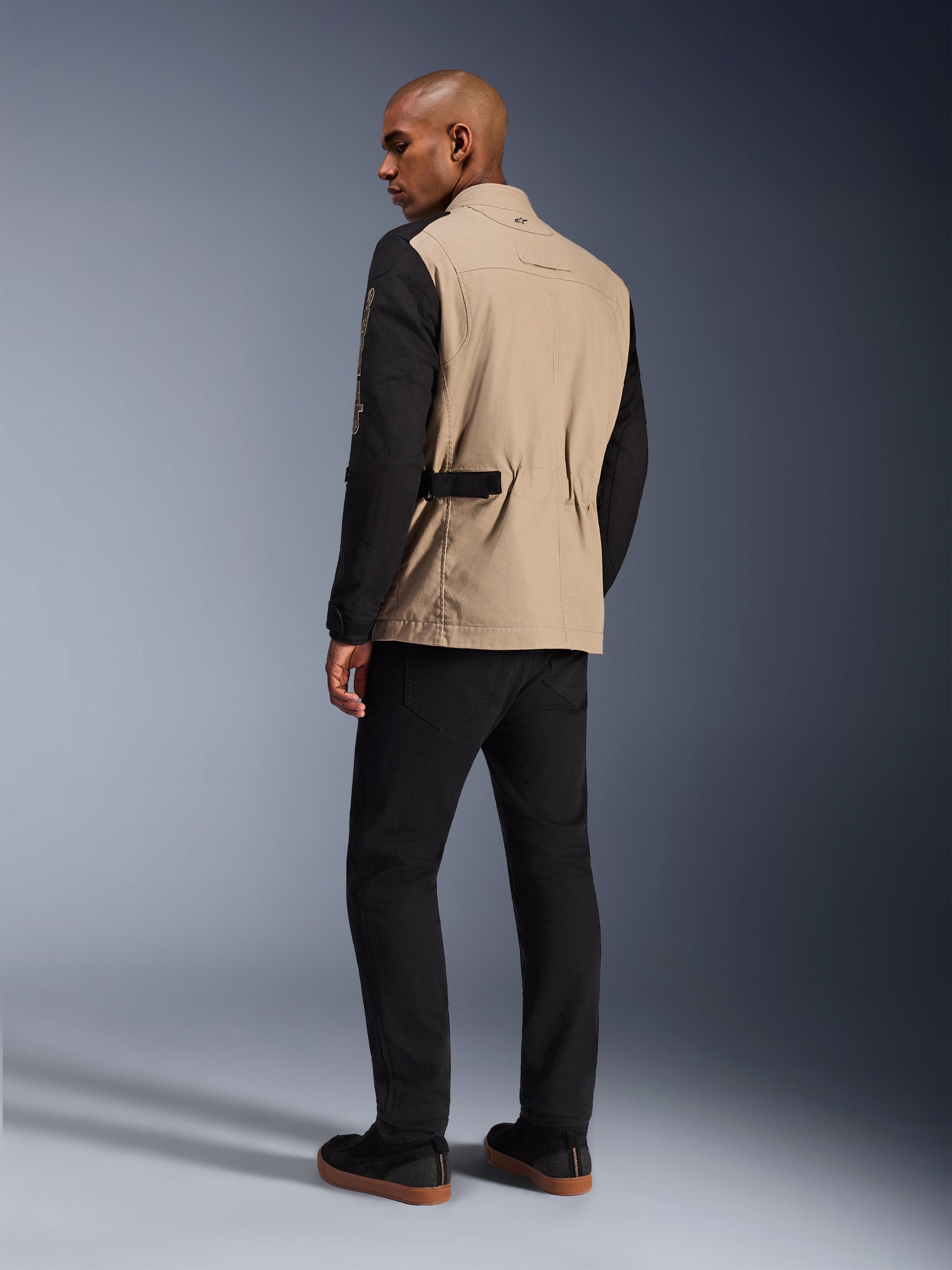 Alpinestars Dice Jas, urban motorjas, Aluminum Black met beige romp en zwarte mouwen, achteraanzicht op model, robuust op werkkleding geïnspireerd ontwerp met tailleverstellers, gecombineerd met een zwarte motorbroek.