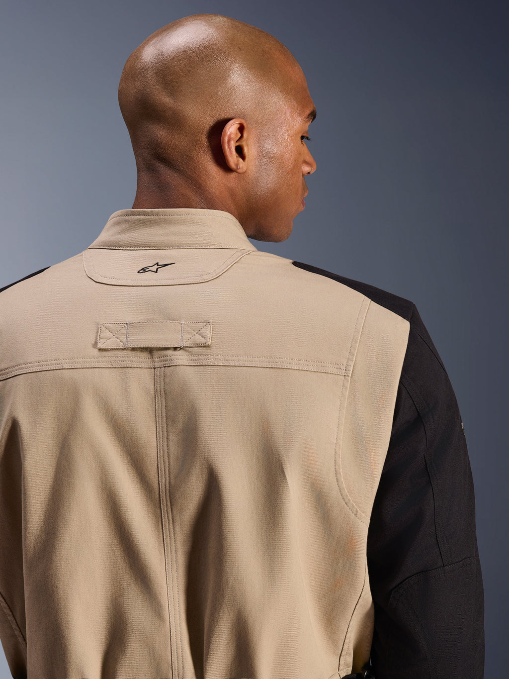 Alpinestars Dice Jacket, stedelijke motorjas, beige en zwart, close-up detail van de achterkant van de kraag en schouder, voorzien van een geborduurd logo, robuuste polykatoen twill constructie, street-ready werkkledingstijl