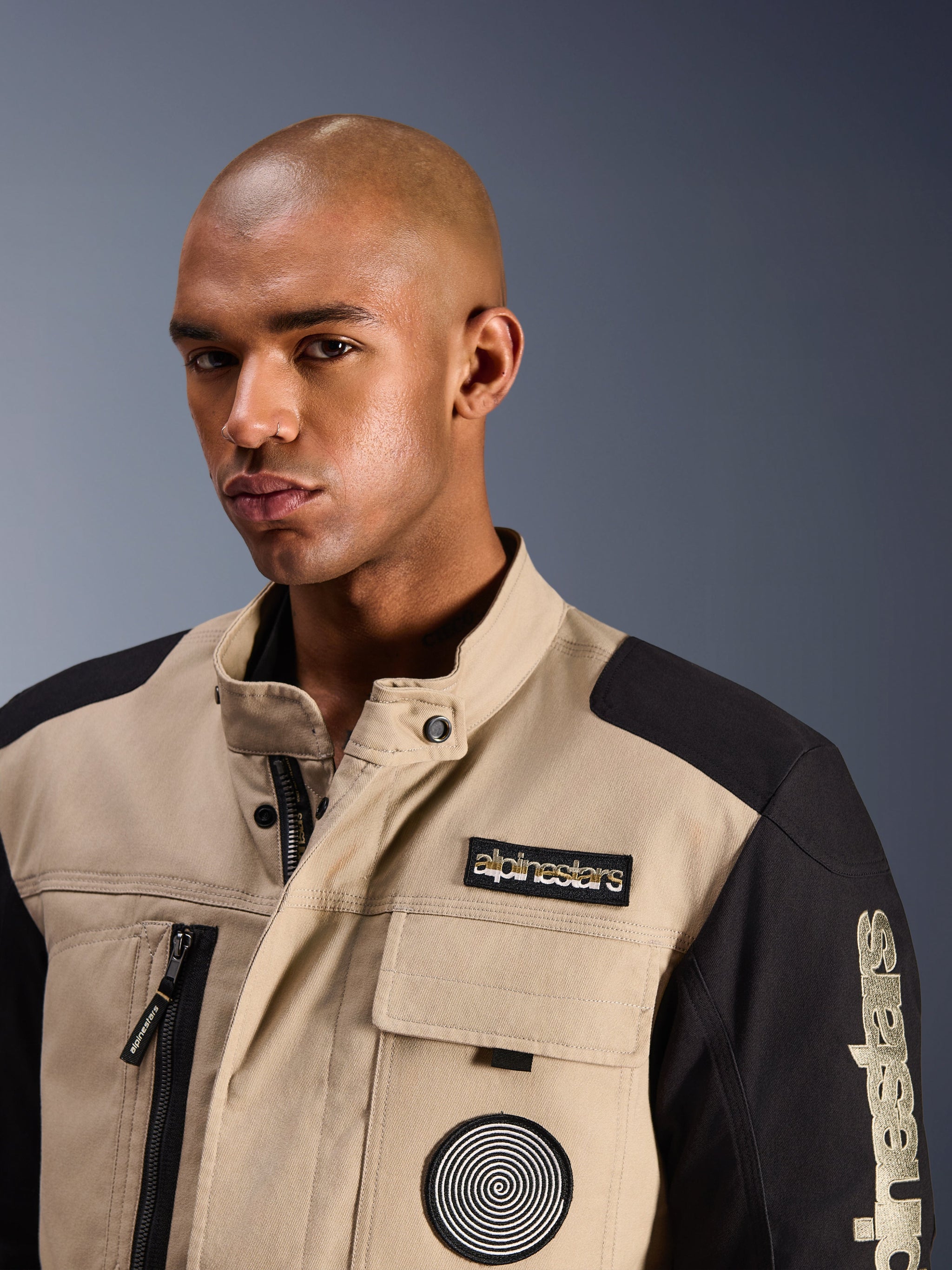 Mannelijk model draagt de Alpinestars Dice Jacket in Aluminum Black, stedelijke motorjas met tan en zwarte kleurvlakken, voorzien van een geborduurde Alpinestars patch op de borst en spiraallogo, robuust op werkkleding geïnspireerd ontwerp met een hoge kraag