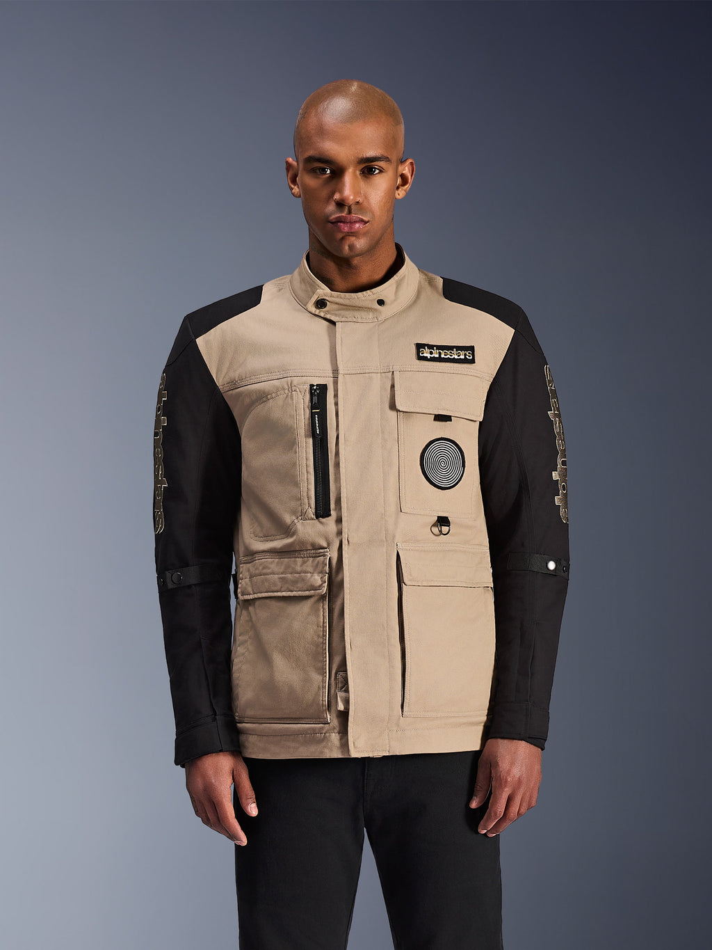 Alpinestars Dice Jacket, stedelijke motorjas, beige en zwart, robuuste werkkleding-esthetiek met meerdere functionele zakken en patches op de borst, hoge kraag met knoopsluiting, opvallende merkdetails op de mouwen, gedragen door een mannelijk model