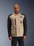 Alpinestars Dice Jacket, stedelijke motorjas, beige en zwart, robuuste werkkleding-esthetiek met meerdere functionele zakken en patches op de borst, hoge kraag met knoopsluiting, opvallende merkdetails op de mouwen, gedragen door een mannelijk model