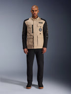 Alpinestars Dice Jacket, stedelijke motorjas, aluminium beige romp met zwarte mouwen, op werkkleding geïnspireerd silhouet met vier praktische zakken aan de voorzijde, gedragen met een zwarte broek en zwarte hoge sneakers, robuuste straatwaardige motoruitrusting.