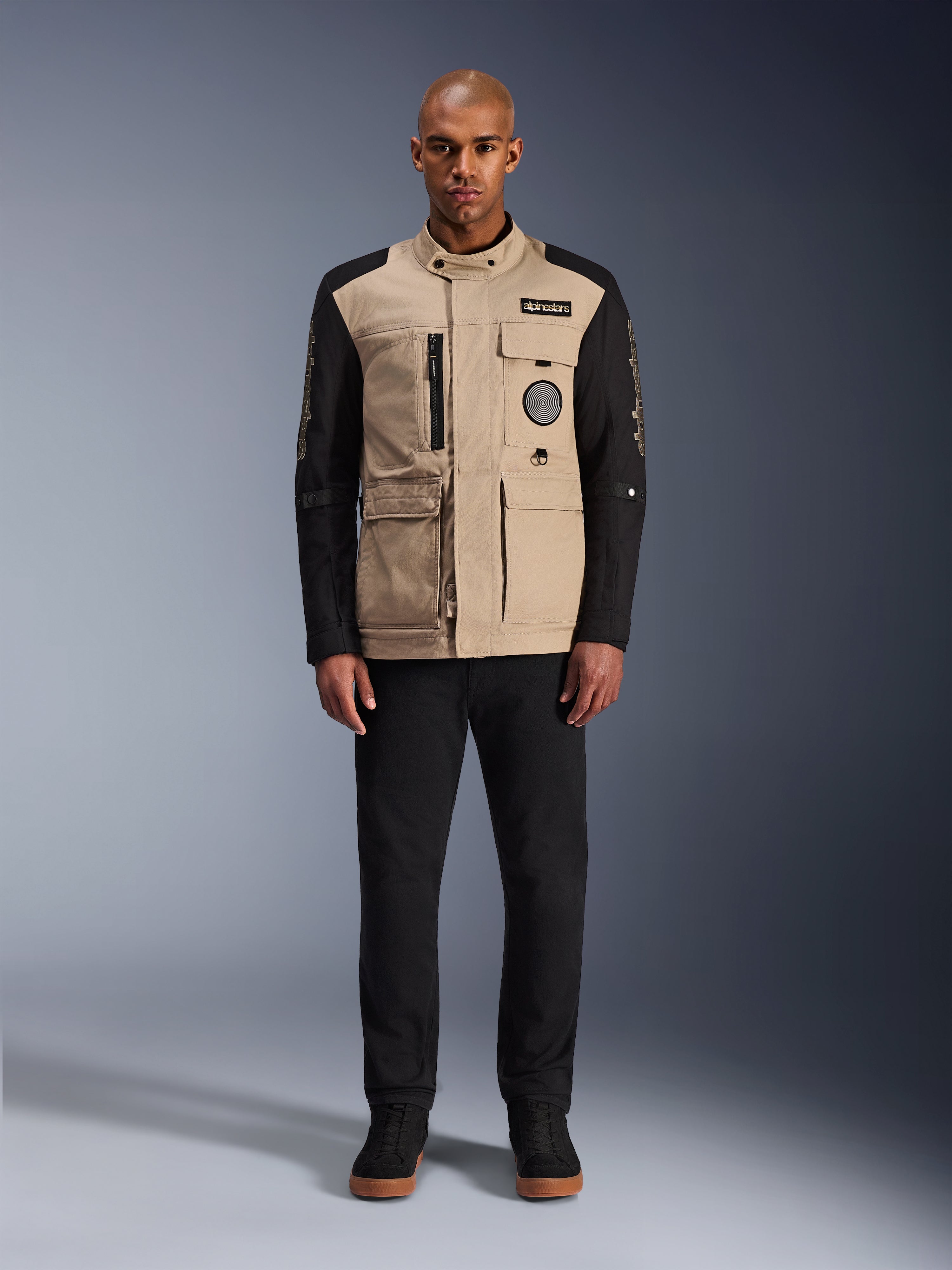 Alpinestars Dice Jacket, stedelijke motorjas, aluminium beige romp met zwarte mouwen, op werkkleding geïnspireerd silhouet met vier praktische zakken aan de voorzijde, gedragen met een zwarte broek en zwarte hoge sneakers, robuuste straatwaardige motoruitrusting.