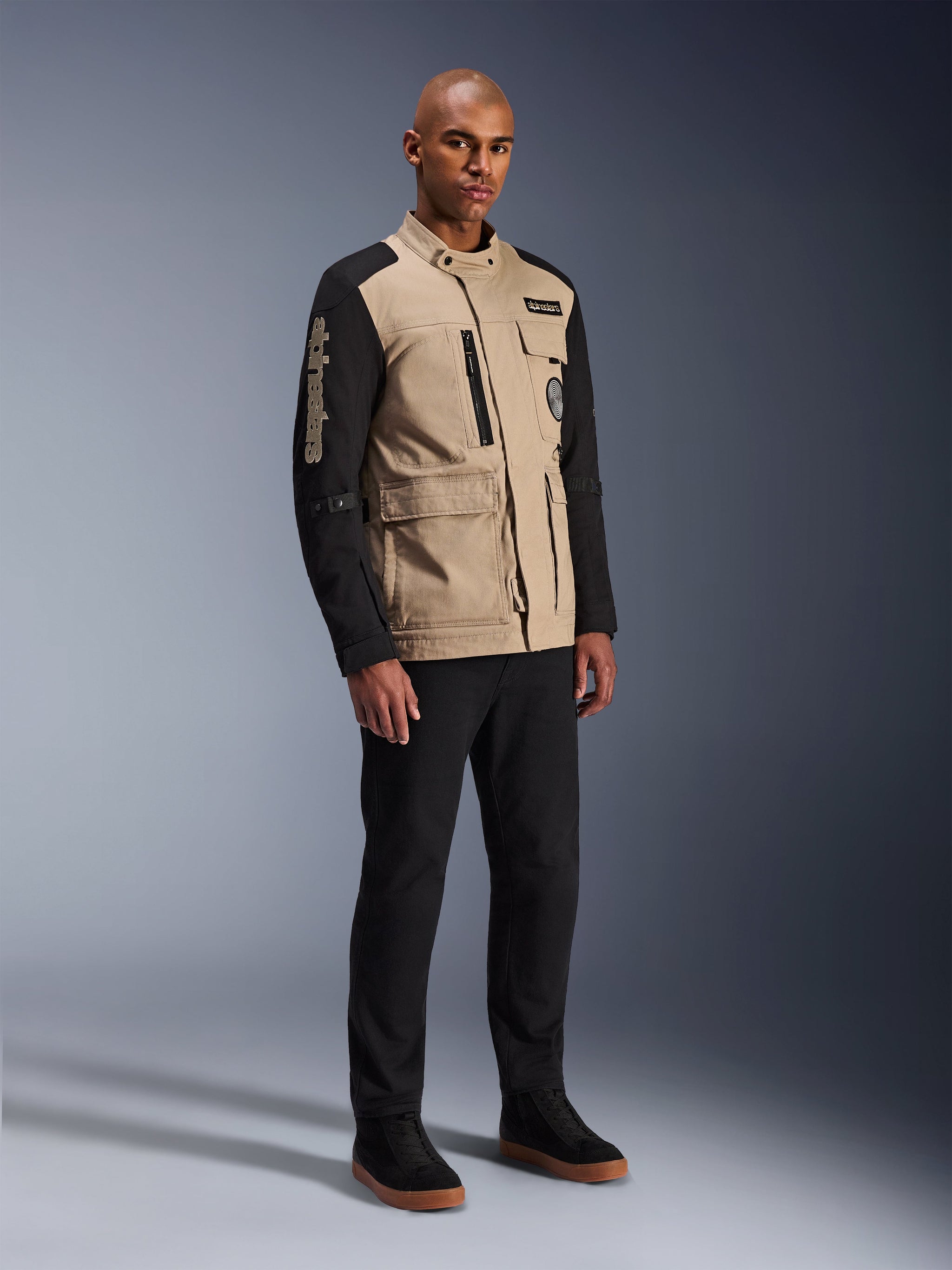 Alpinestars Dice Jas, stedelijke motorjas, Aluminum Black en Tan, tweekleurig ontwerp met zwarte mouwen en een khaki torso, gedragen door een model met zwarte broek en zwarte sneakers, op werkkleding geïnspireerde esthetiek met functionele zakken en geborduurd logo op de mouw.