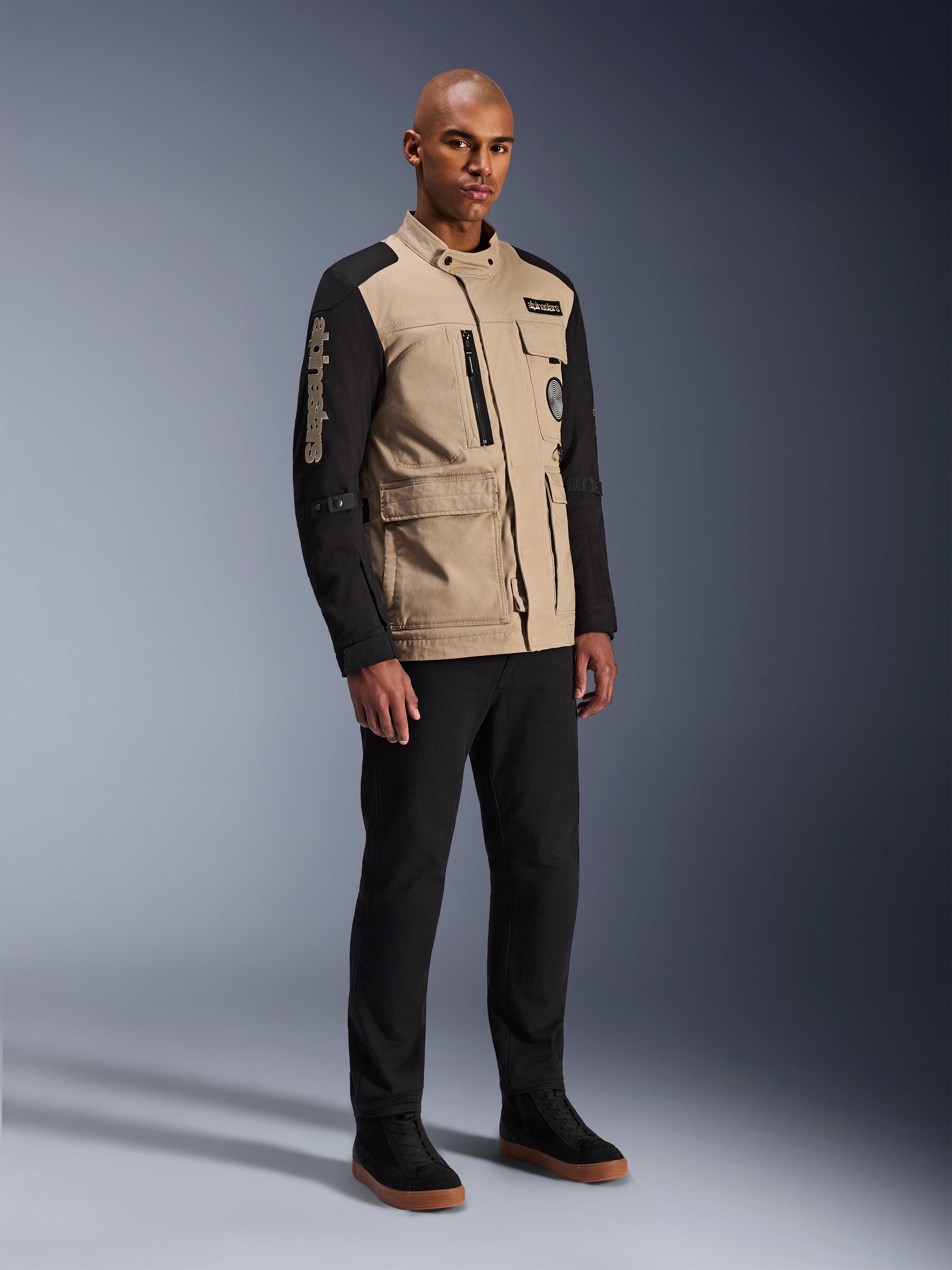 Alpinestars Dice Jas, stedelijke motorjas, Aluminum Black en Tan, tweekleurig ontwerp met zwarte mouwen en een khaki torso, gedragen door een model met zwarte broek en zwarte sneakers, op werkkleding geïnspireerde esthetiek met functionele zakken en geborduurd logo op de mouw.