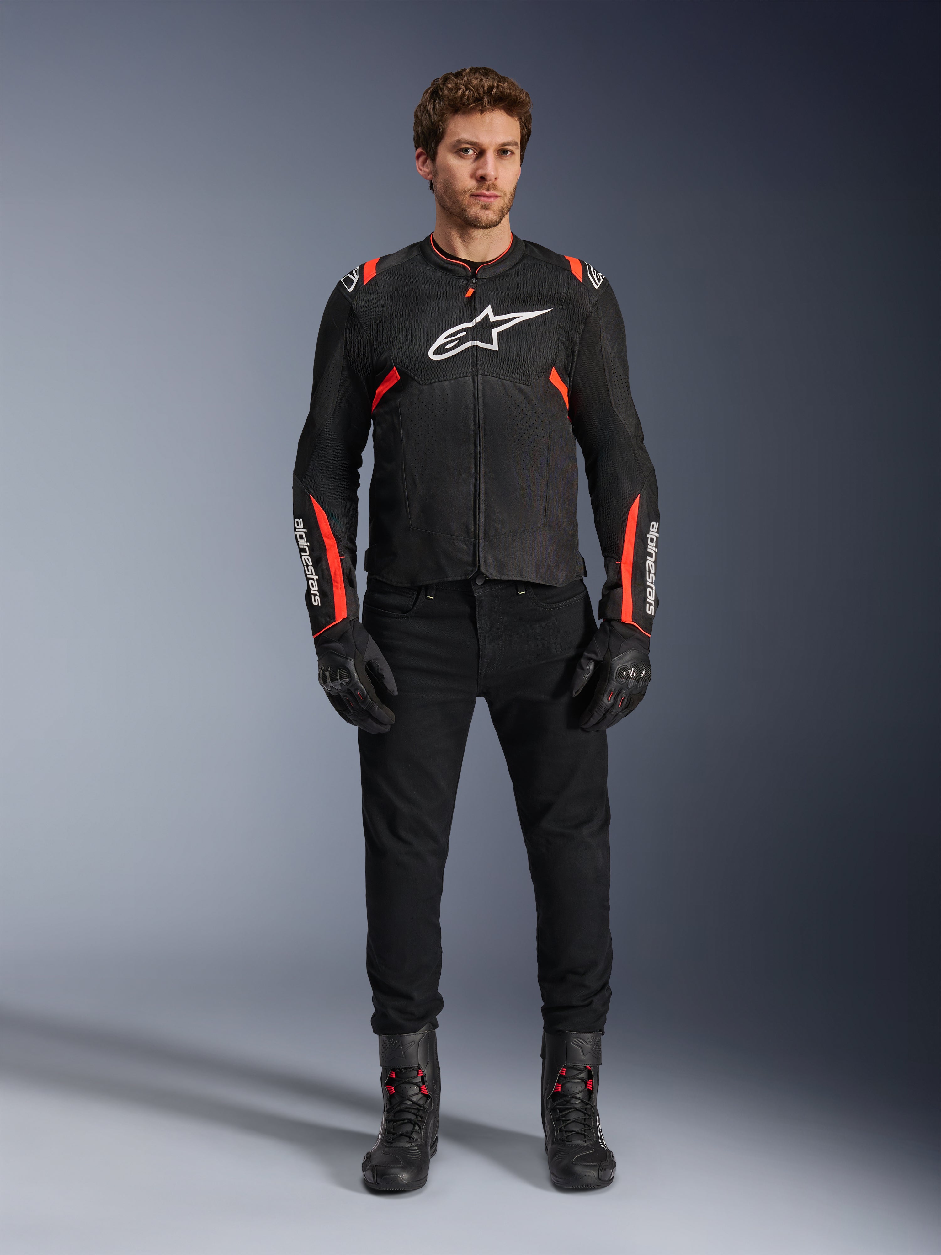 Mannelijk model draagt Alpinestars T-SPS Air v2 jas, zwarte textiele motorjas met Red Fluo accenten, wit logo op de borst, gedragen met zwarte broek, handschoenen en laarzen, urban sport rijstijl