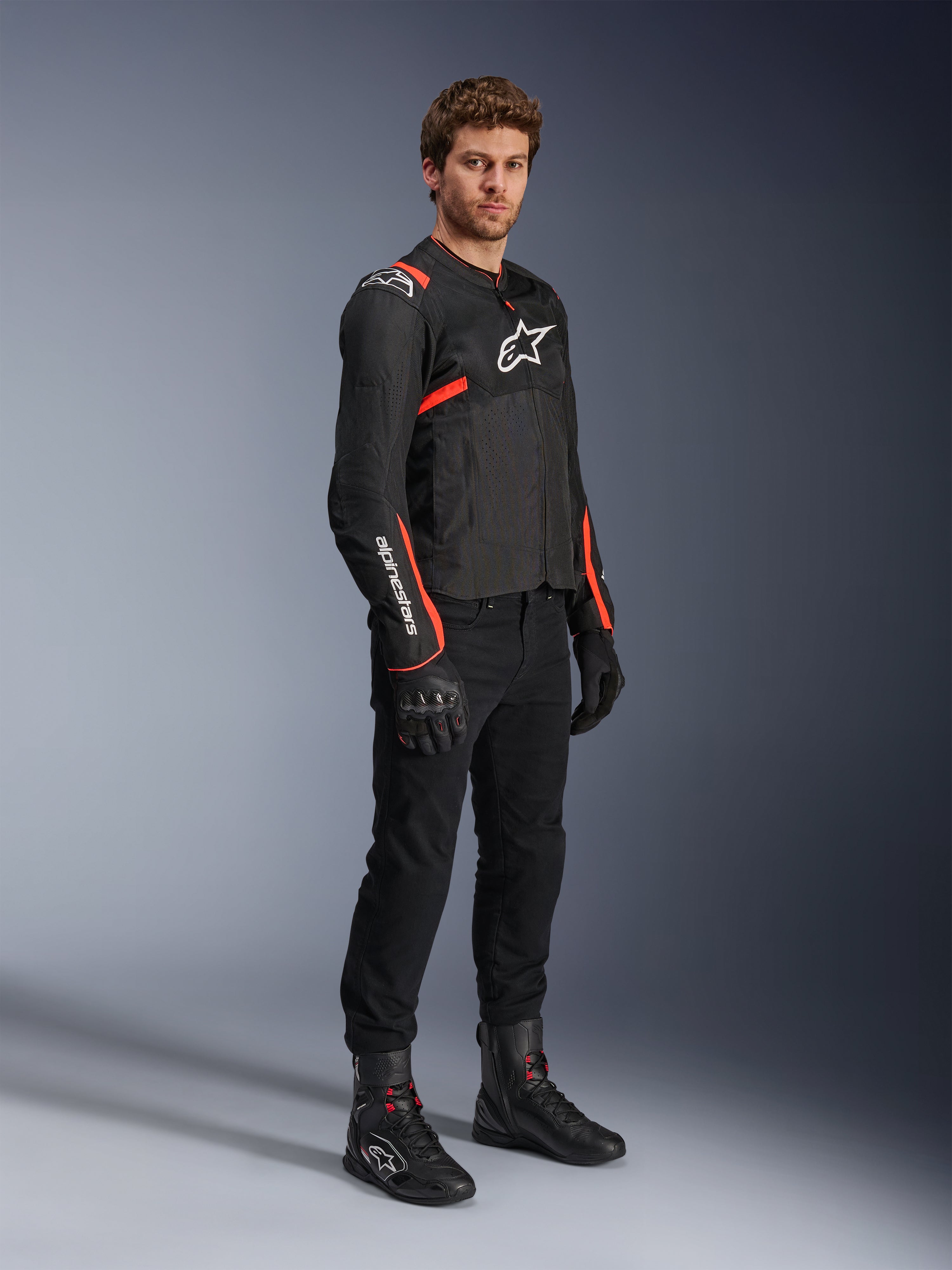 Mannelijk model in Alpinestars T-Sps Air v2 Jas, zwart met Red Fluo accenten, gecombineerd met een zwarte motorbroek, zwarte handschoenen en laarzen, sportief ontwerp met mesh-panelen en witte Alpinestars-logo's