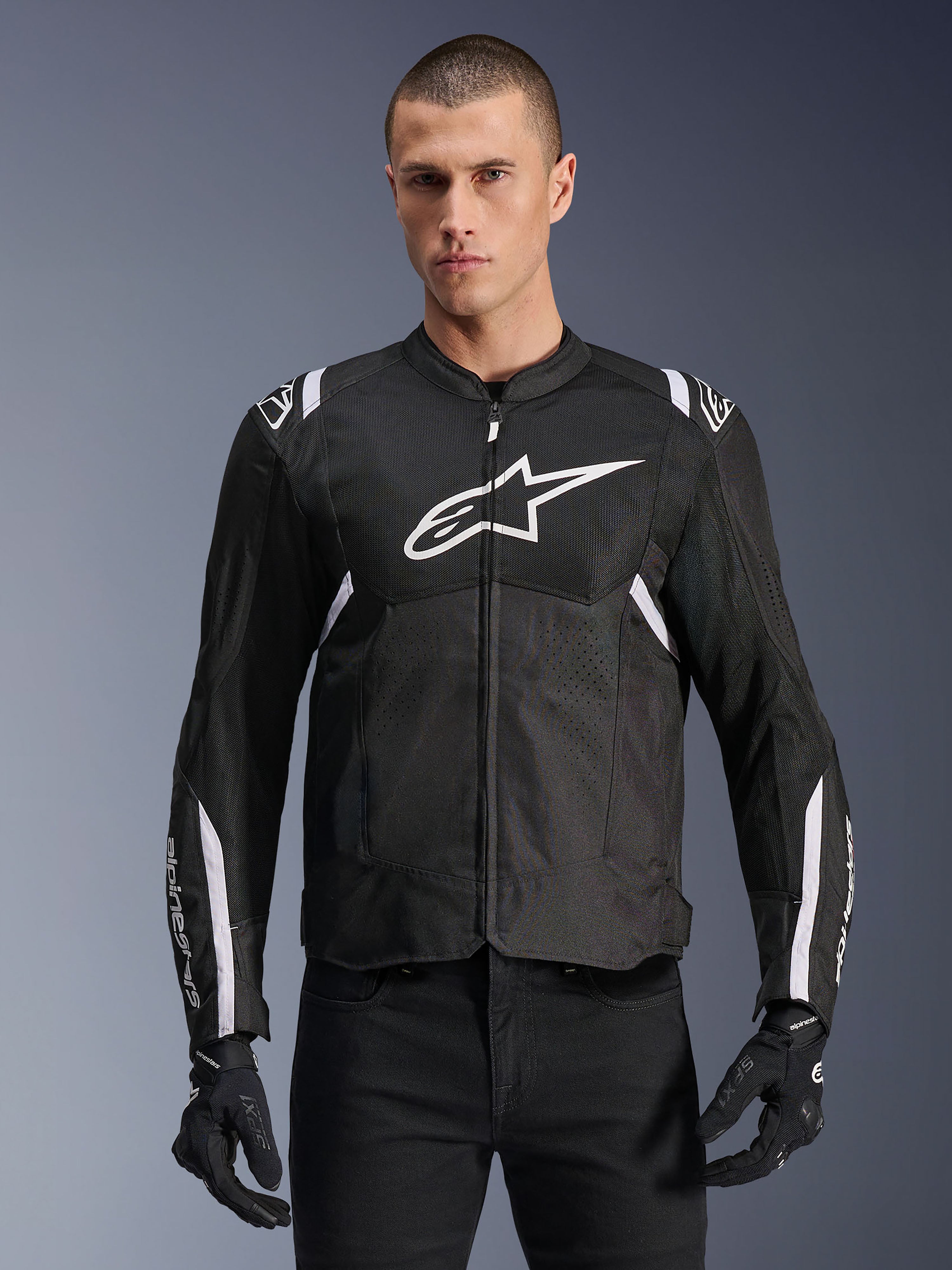 Alpinestars T-SPS Air V2 motorjas, textiele motorjas, zwart met witte accenten, groot wit logo op de borst, geperforeerd mesh-ontwerp voor ventilatie, gedragen met een zwarte broek en handschoenen