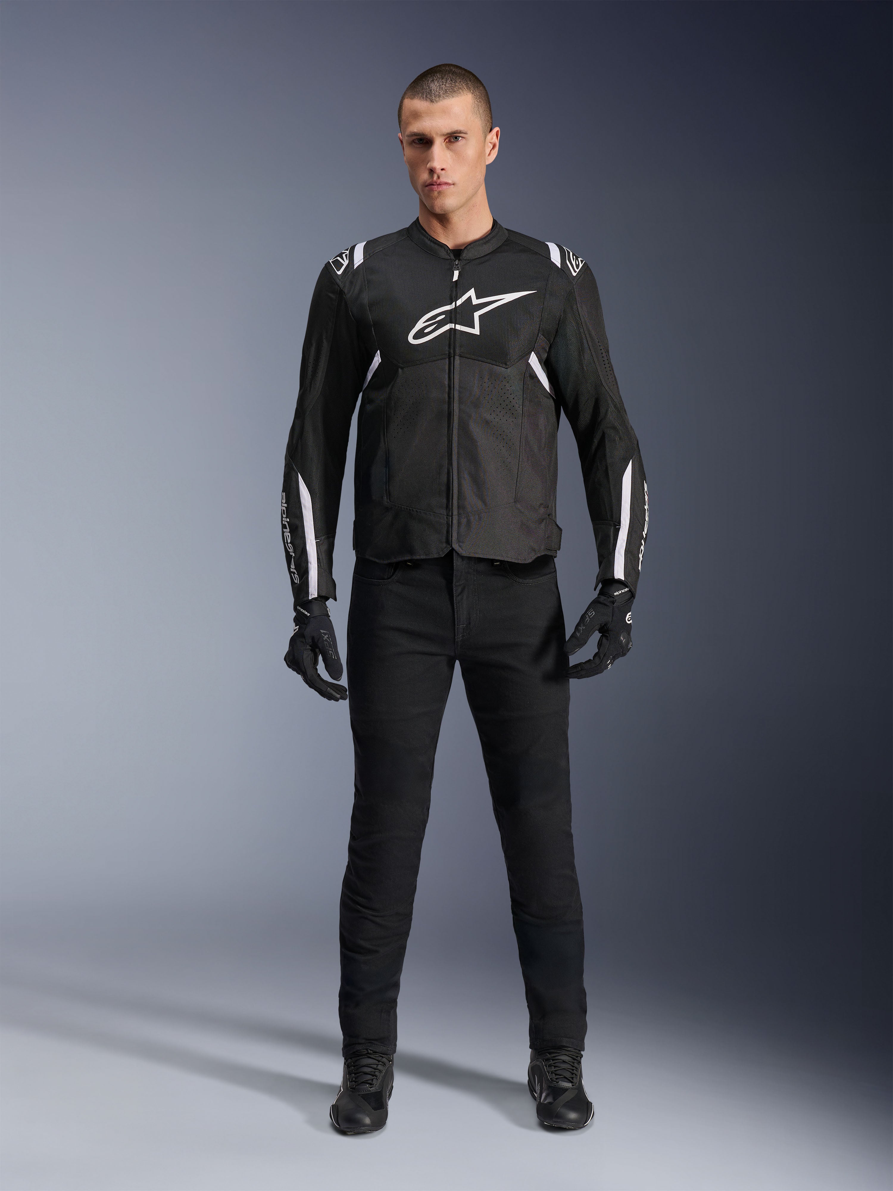 Alpinestars T-Sps Air V2 Jas, zwart-witte textiele motorjas, met een groot wit logo op de borst en mesh-panelen, gedragen door een model met zwarte uitrusting voor sportief stedelijk rijden