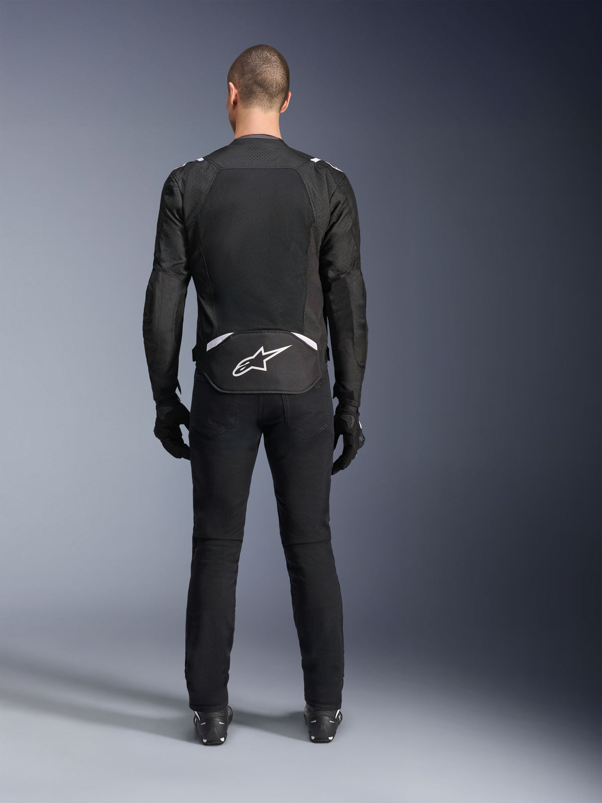 Alpinestars T-Sps Air V2 Jas, achteraanzicht, zwart-witte textiele motorjas, voorzien van een groot mesh rugpaneel voor maximale luchtstroom en wit Astars-logo op de onderrug, gedragen met een zwarte motorbroek en handschoenen