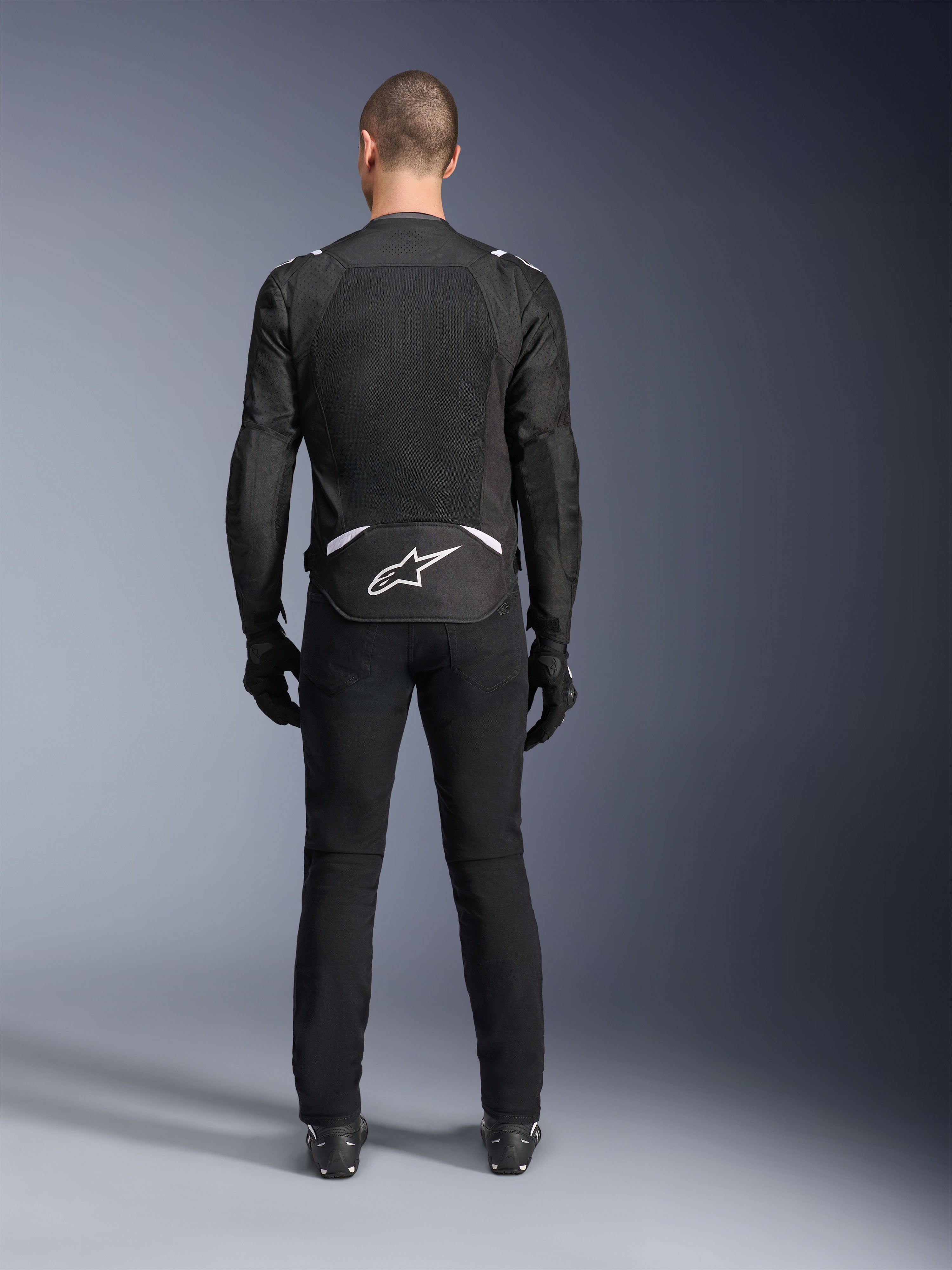 Alpinestars T-Sps Air V2 Jas, achteraanzicht, zwart-witte textiele motorjas, voorzien van een groot mesh rugpaneel voor maximale luchtstroom en wit Astars-logo op de onderrug, gedragen met een zwarte motorbroek en handschoenen