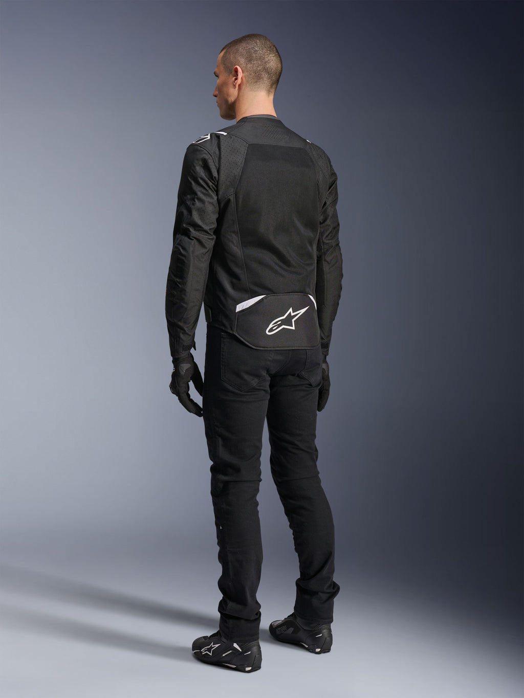 Alpinestars T-Sps Air V2 Jas, textiele motorjas, zwart, achteraanzicht met groot mesh ventilatiepaneel en wit Alpinestars-logo op de onderrug, gecombineerd met een zwarte motorbroek en handschoenen voor een sportieve urban look.
