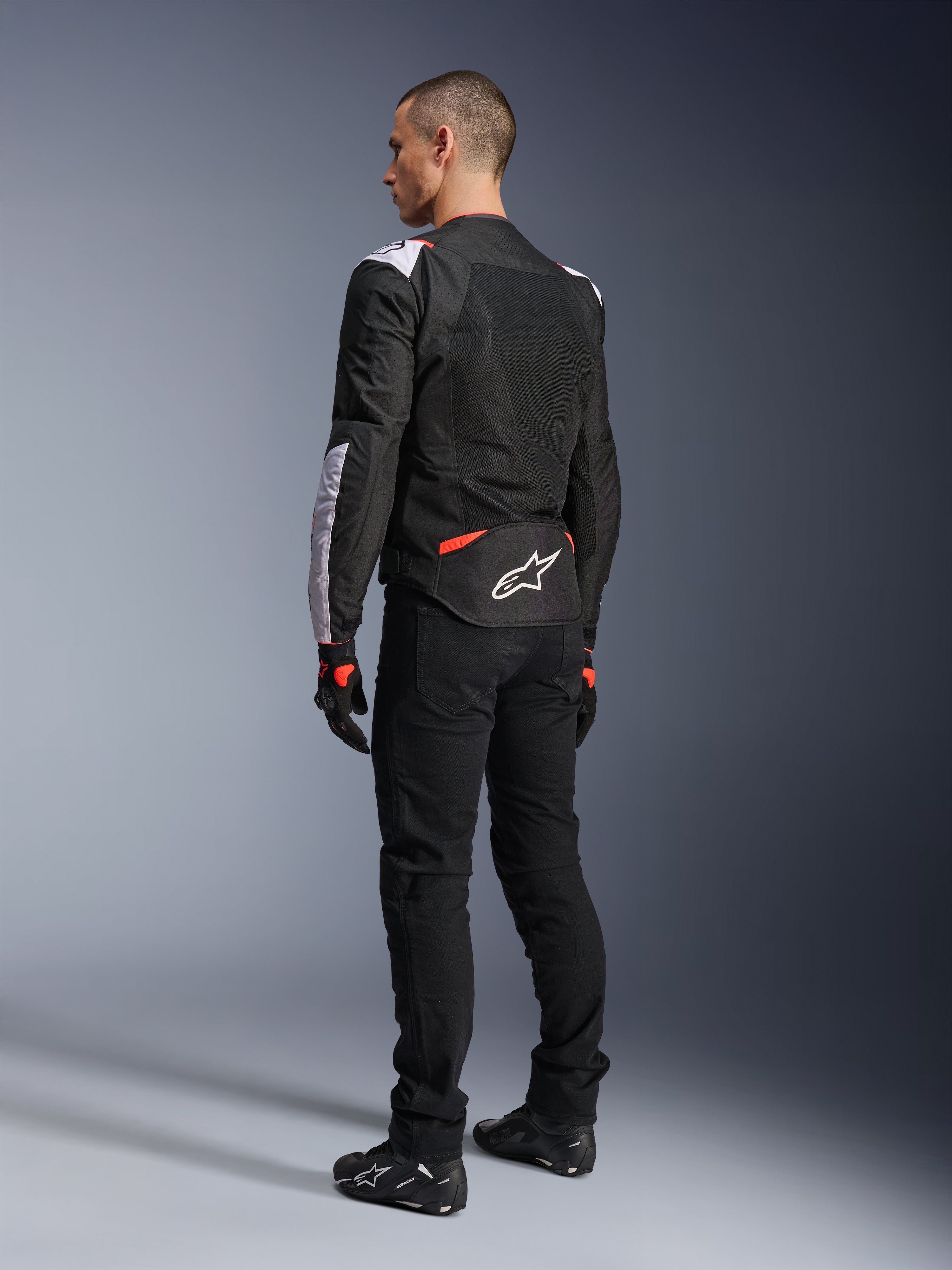 Alpinestars T-Sps Air V2 Jas, textiel motorjas, zwart, wit en fluorood, achteraanzicht op model, mesh-panelen voor ventilatie, wit Astars-logo op de onderrug, gecombineerd met zwarte motorbroek en handschoenen