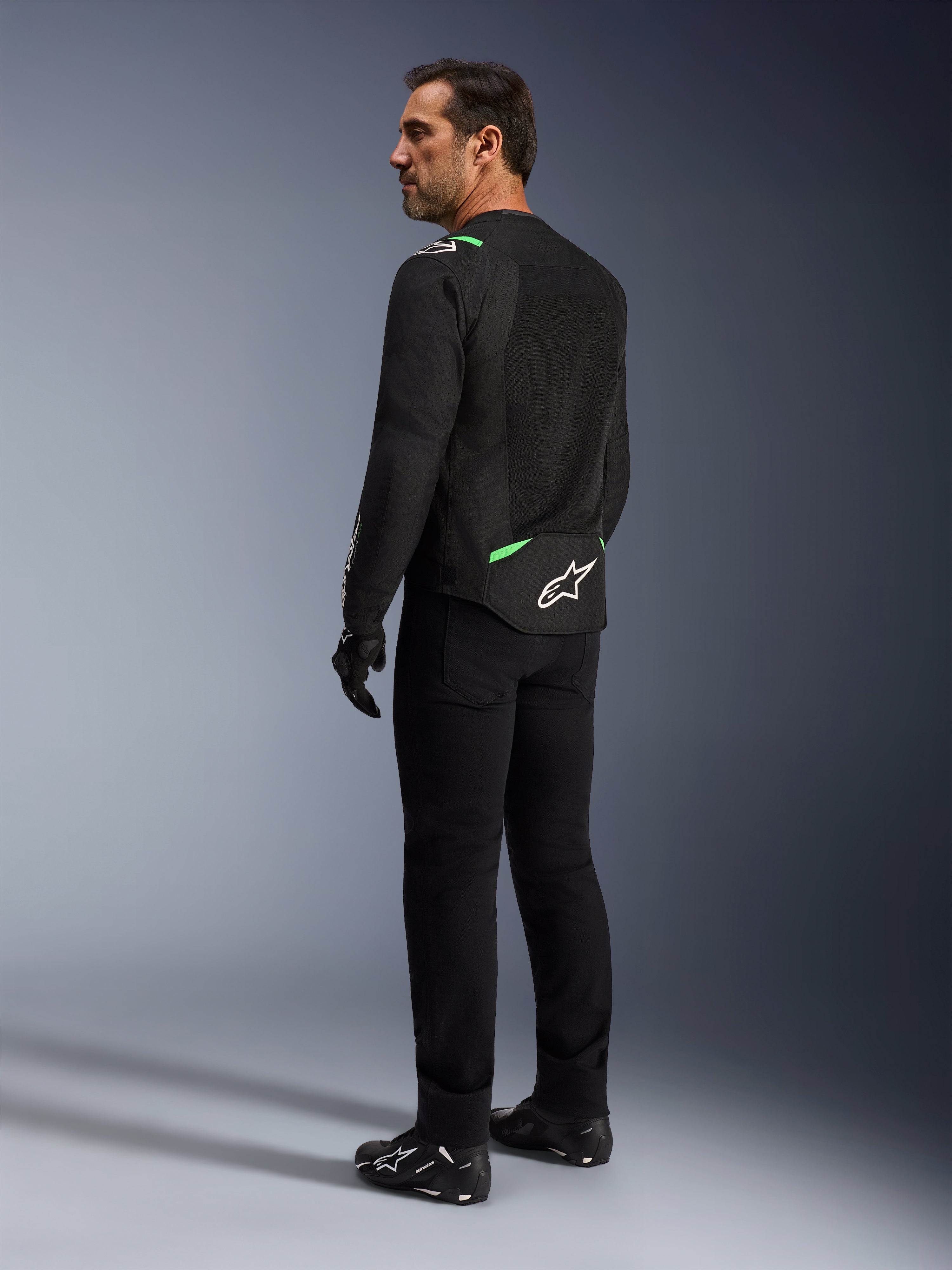 Alpinestars T-SPS Air V2 Jas in Black Fluo Green, man getoond vanuit rug- en zijprofiel, gecombineerd met zwarte motorbroek en motorschoenen, voorzien van geventileerde tech mesh panelen, felgroene accenten op de schouders en onderrug, wit sterlogo op de achterzijde.