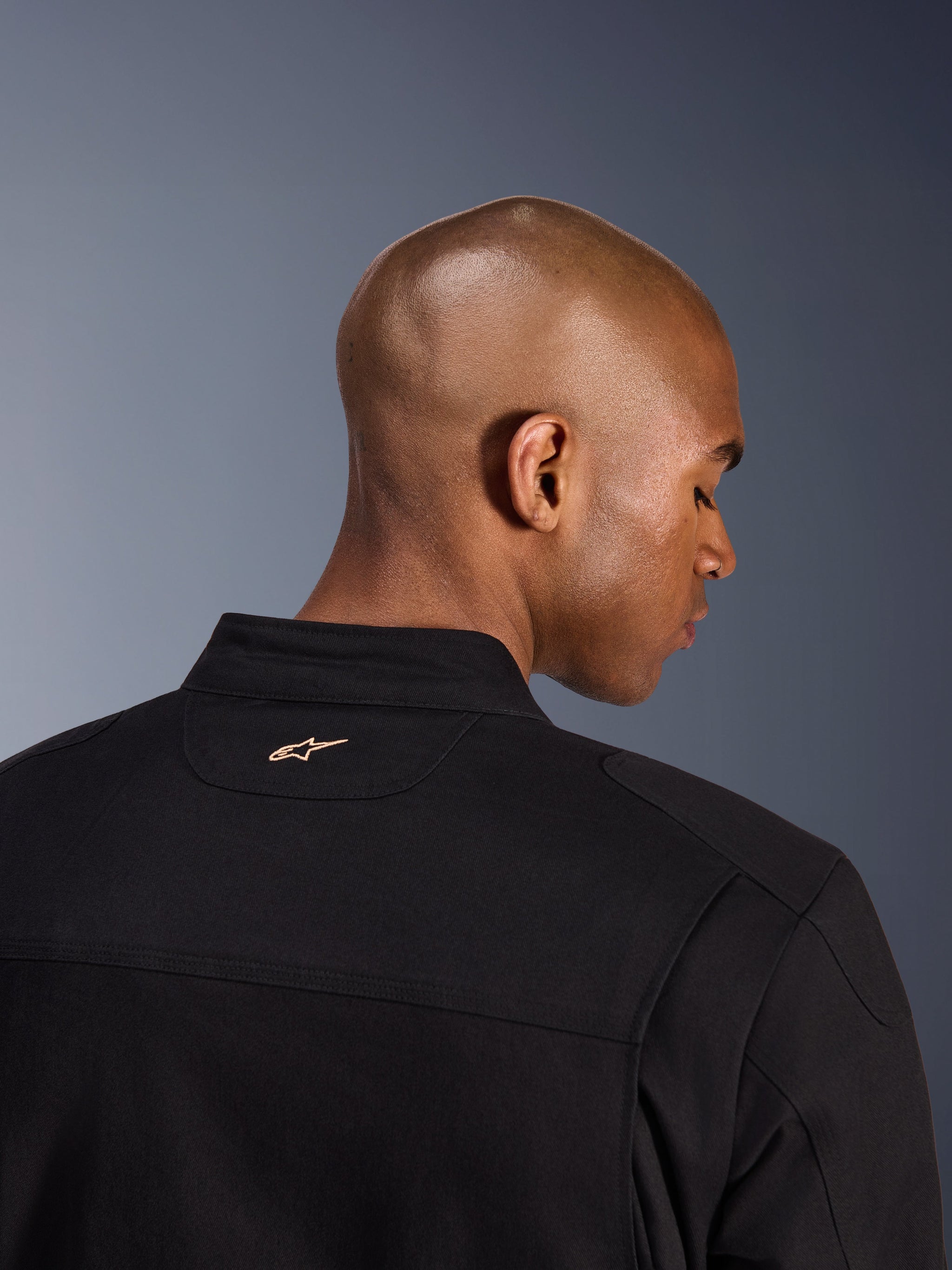 Alpinestars Bruiser Jacket, zwart, detailopname van de achterkant van de kraag en schouders, met een klein lichtbruin geborduurd Alpinestars-logo op de nek, stedelijke motorjas met workwear-esthetiek