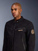 Mannelijk model draagt Alpinestars Bruiser Jacket, zwarte urban motorjas van polycotton twill, voorzien van zakken met ritssluiting, goud geborduurd logo op de borst en Oscar-spiraalpatch op de mouw, moderne stijl voor woon-werkverkeer