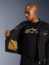 Alpinestars Bruiser Jacket, zwarte urban motorjas, close-up van de open binnenkant met goudkleurige mesh zak en 'Alpinestars Protects' branding, gedragen over een zwart t-shirt met logo, textiele motorkleding met werkkleding-esthetiek