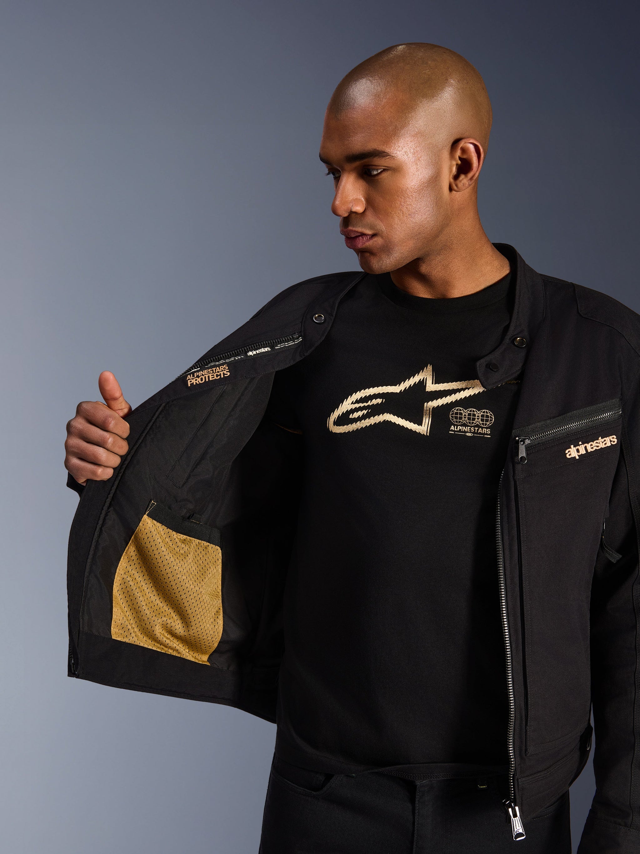 Alpinestars Bruiser Jacket, zwarte urban motorjas, close-up van de open binnenkant met goudkleurige mesh zak en 'Alpinestars Protects' branding, gedragen over een zwart t-shirt met logo, textiele motorkleding met werkkleding-esthetiek