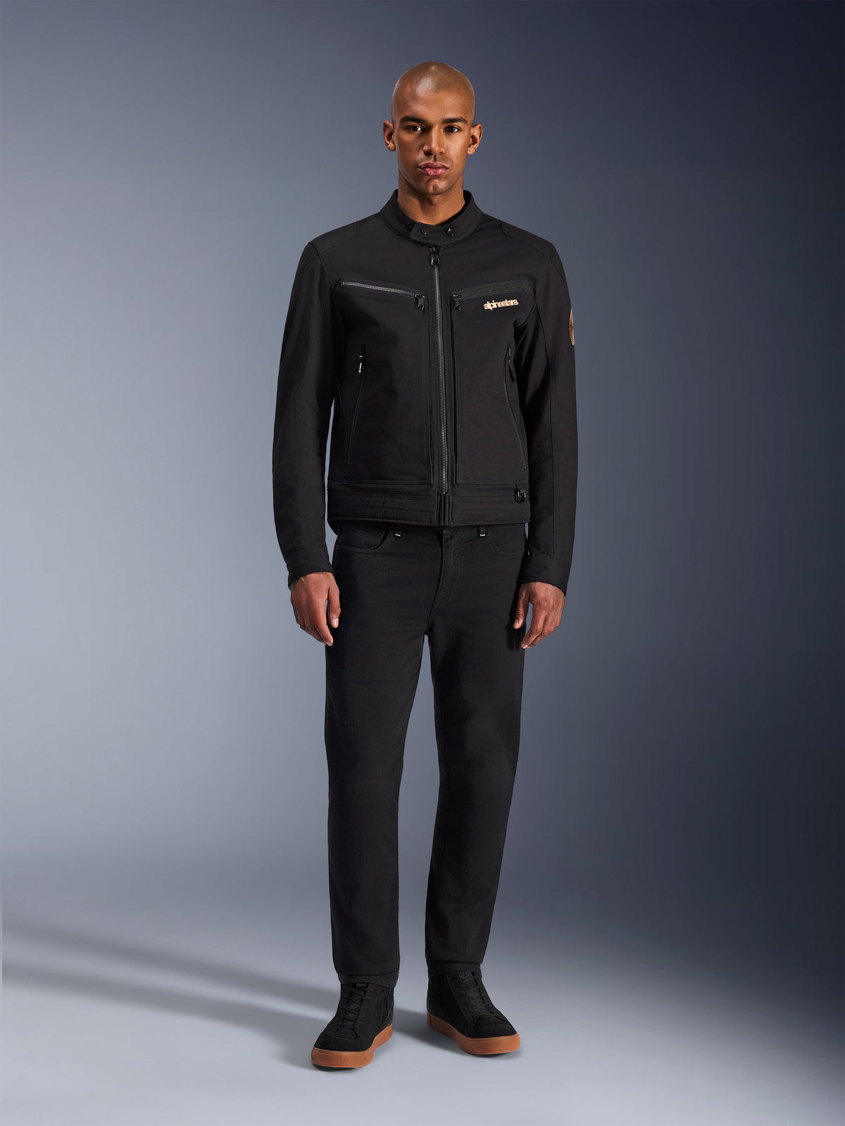 Alpinestars Bruiser Jas, zwarte urban motorjas, gedragen door model met zwarte broek en hoge sneakers, vooraanzicht met borstzakken met ritssluiting en geborduurd logo, klassiek op werkkleding geïnspireerd ontwerp voor dagelijks woon-werkverkeer