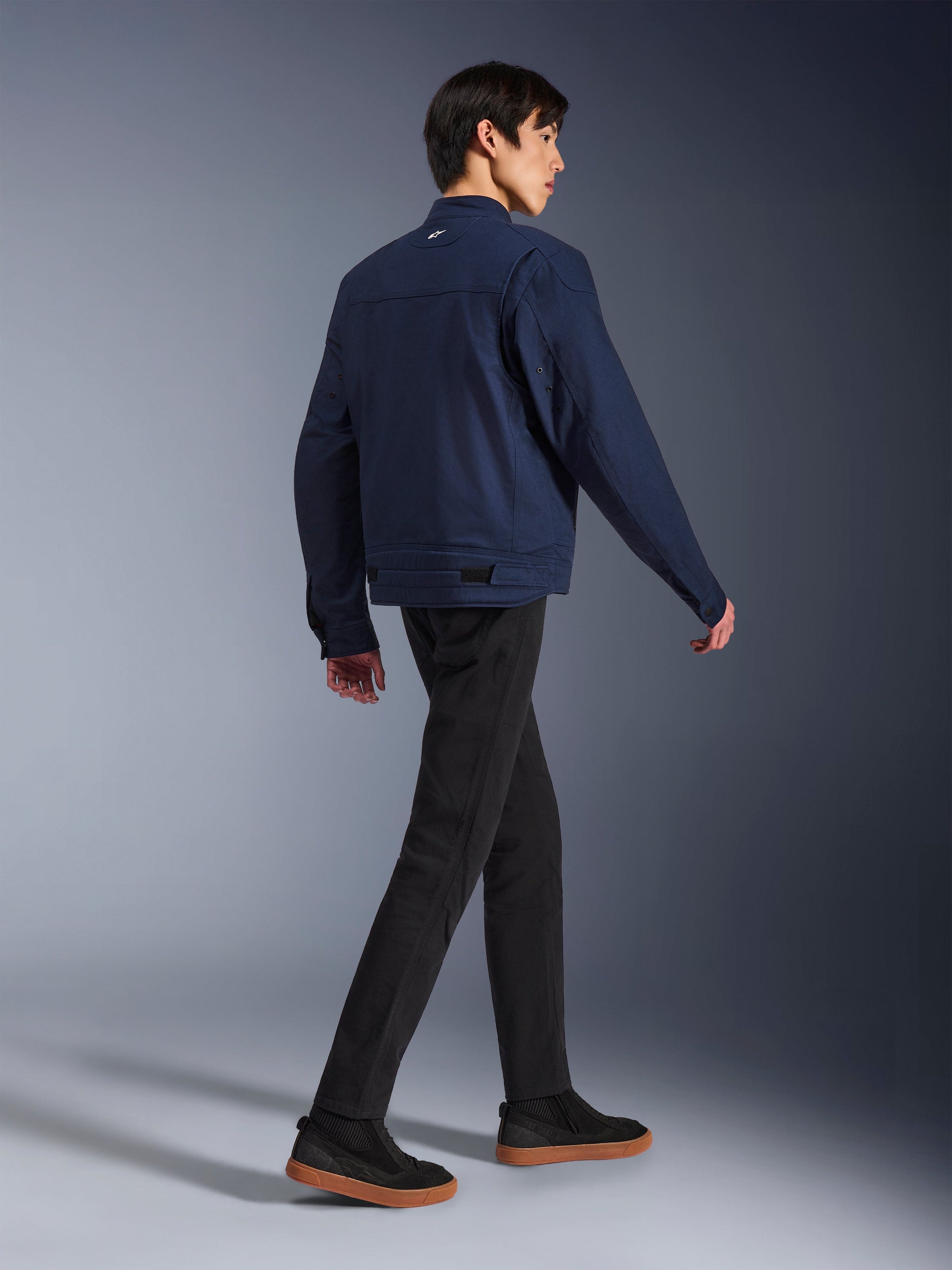 Alpinestars Bruiser Jacket in Dark Blue, urban textile motorcycle jacket, rear view on model walking, paired with black pants and gum-soled sneakers, featuring workwear-inspired design and adjustable waist tabs