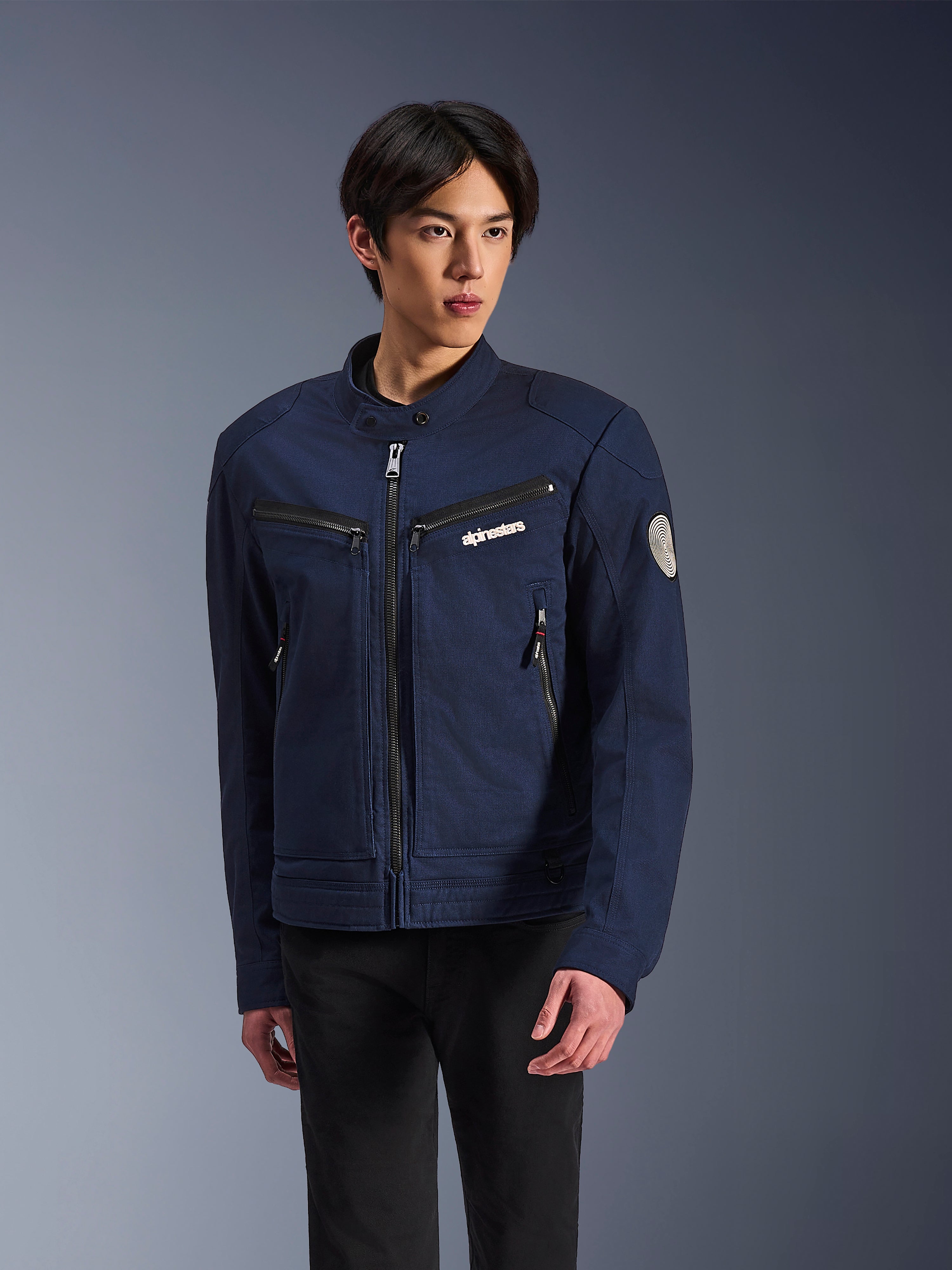 Alpinestars Bruiser Jacket, donkerblauwe textiele motorjas, gedragen door een man met zwarte broek, voorzien van ritszakken, wit Alpinestars-logo op de borst en ronde Oscar-spiraalpatch op de mouw, urban streetwear-stijl voor motorrijders