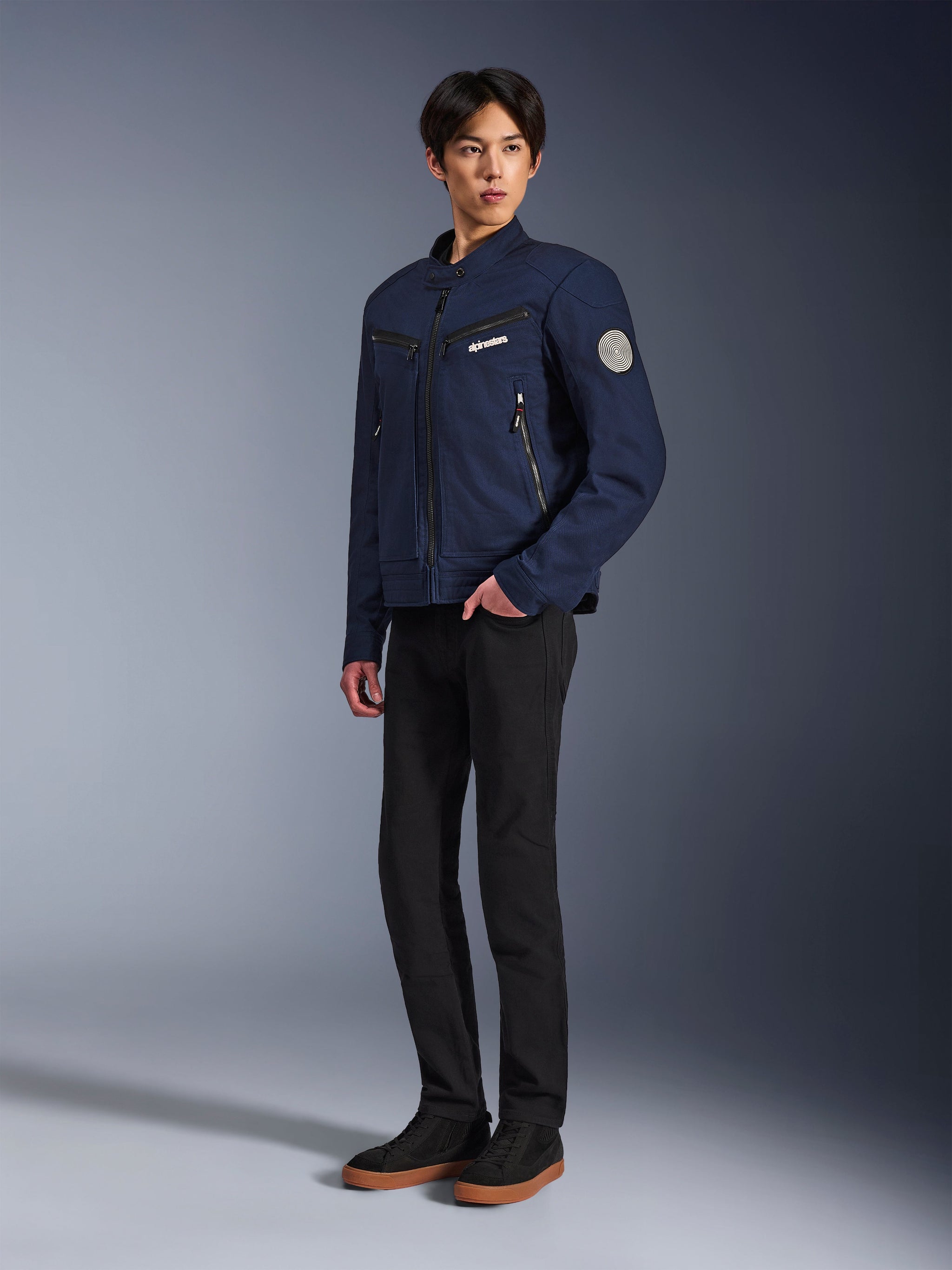 Alpinestars Bruiser Jacket in Dark Blue, urban textile motorcycle jacket with workwear-inspired design, featuring zippered chest pockets and Oscar spiral sleeve patch, worn with black pants and casual sneakers.