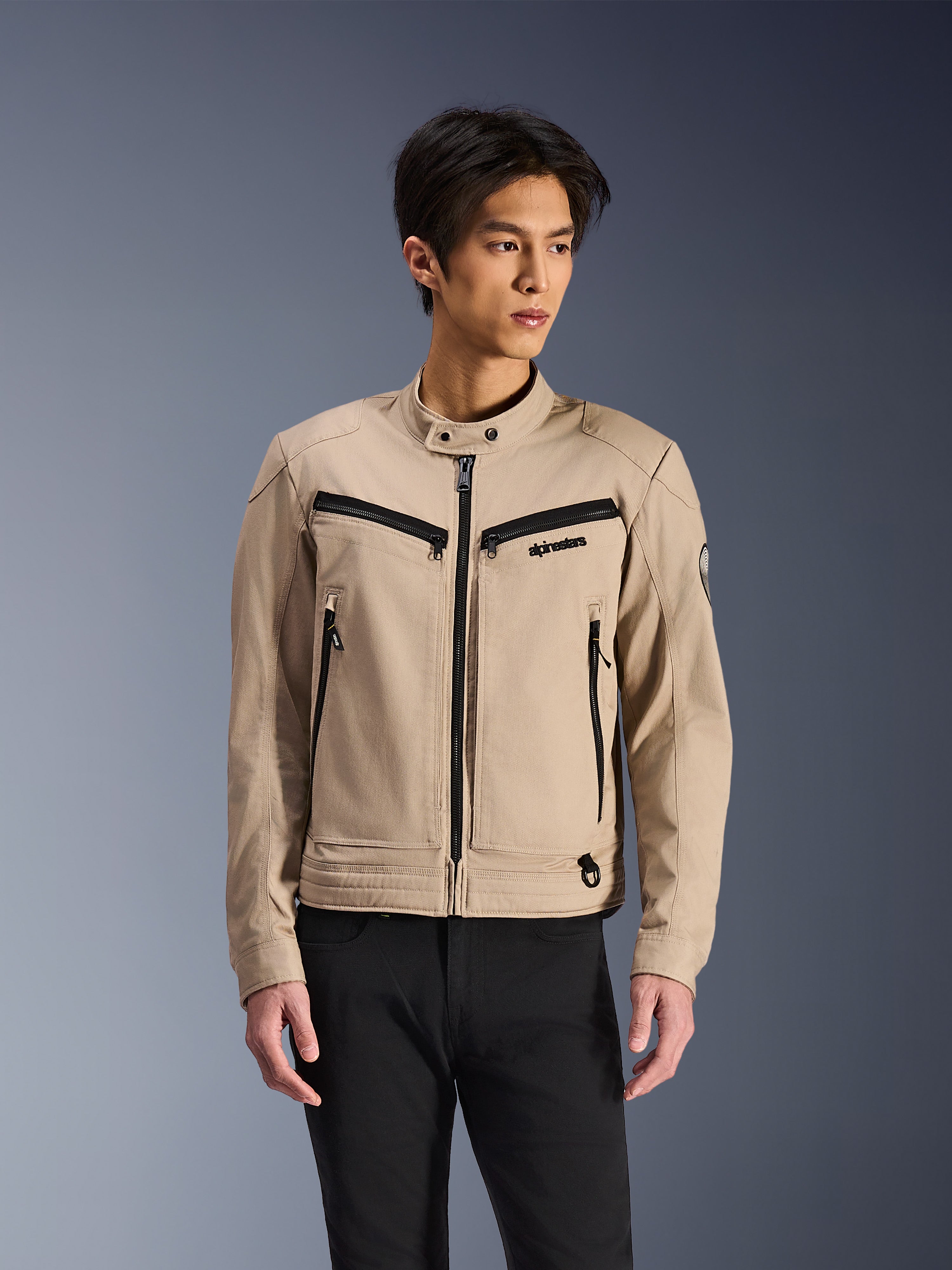 Alpinestars Bruiser Jacket, beige motorjas gedragen door een mannelijk model, voorzien van zwarte ritsdetails op de borst en zijzakken, hoge kraag met drukknopen, op werkkleding geïnspireerde urban motorkleding, gecombineerd met een zwarte broek