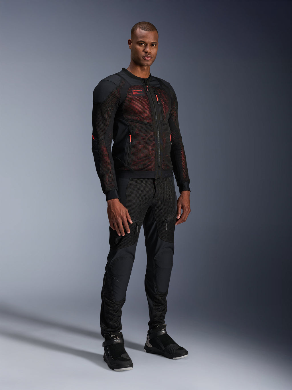 Alpinestars Troop-Air Pro Jas in Black Red Fluo, technische gepantserde mesh motorjas gedragen door een mannelijk model met bijpassende zwarte gepantserde broek en hoge motorlaarzen, volledige studio-opname.