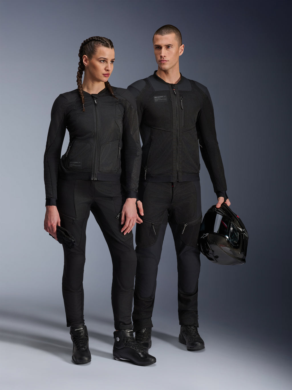 Alpinestars Stella Troop-Air jas, man en vrouw in bijpassende volledig zwarte technische motoruitrusting, voorzien van mesh-constructie voor luchtstroom, zwarte broek en laarzen, man houdt een zwarte helm vast, urban motorkleding