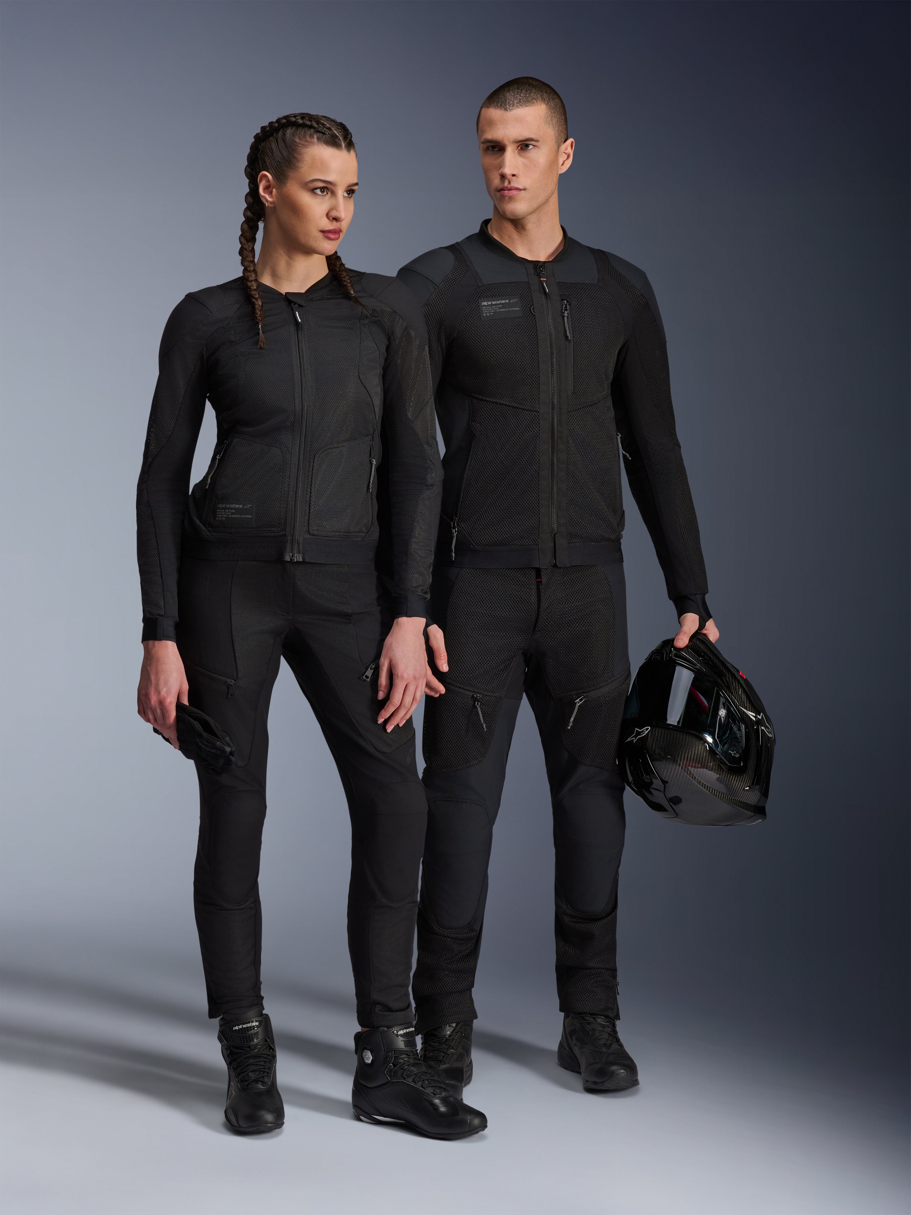 Alpinestars Stella Troop-Air jas, man en vrouw in bijpassende volledig zwarte technische motoruitrusting, voorzien van mesh-constructie voor luchtstroom, zwarte broek en laarzen, man houdt een zwarte helm vast, urban motorkleding