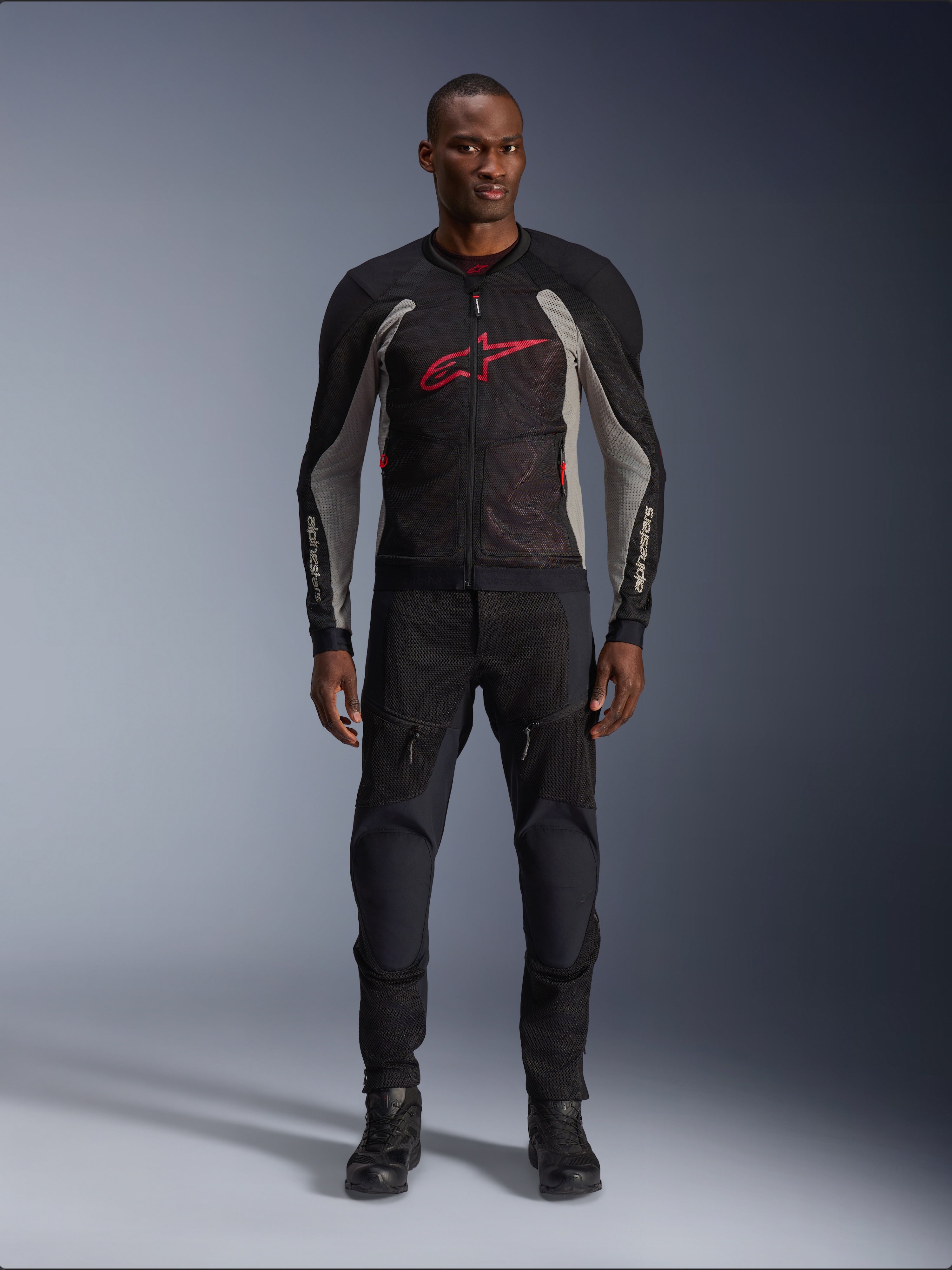 Mannelijk model draagt de Alpinestars Troop-Air Jas in Black Ash Gray en Bright Red, technische mesh motorjas met een opvallend rood logo op de borst, gecombineerd met een zwarte motorbroek en laarzen, stedelijke motorkleding