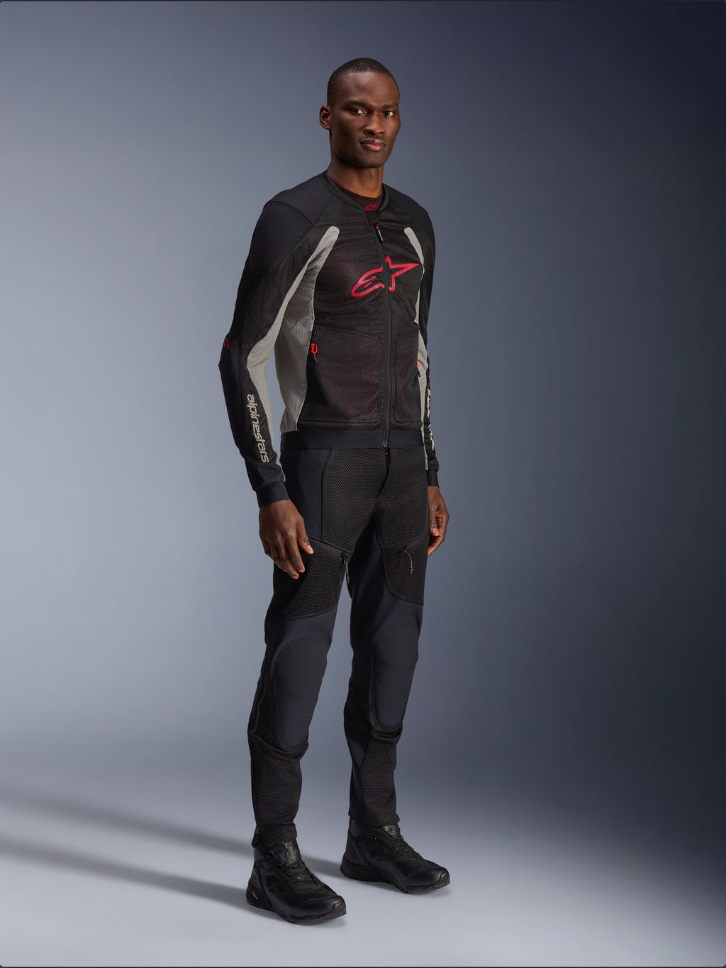 Alpinestars Troop-Air Jas, technische mesh motorjas in zwart, asgrijs en helderrood, gedragen door een model met bijpassende zwarte broek en laarzen, met een rood Alpinestars-logo op de borst, urban contemporary motorkleding