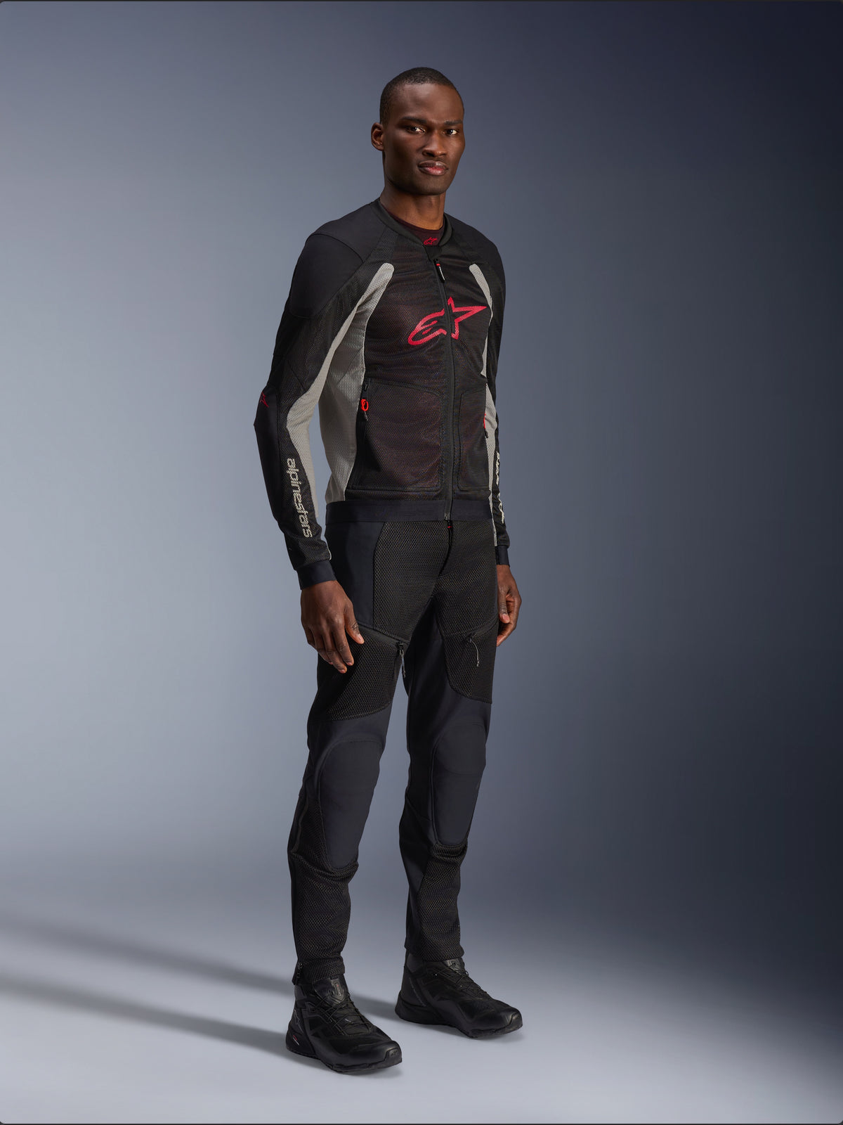 Alpinestars Troop-Air Jas, technische mesh motorjas in zwart, asgrijs en helderrood, gedragen door een model met bijpassende zwarte broek en laarzen, met een rood Alpinestars-logo op de borst, urban contemporary motorkleding