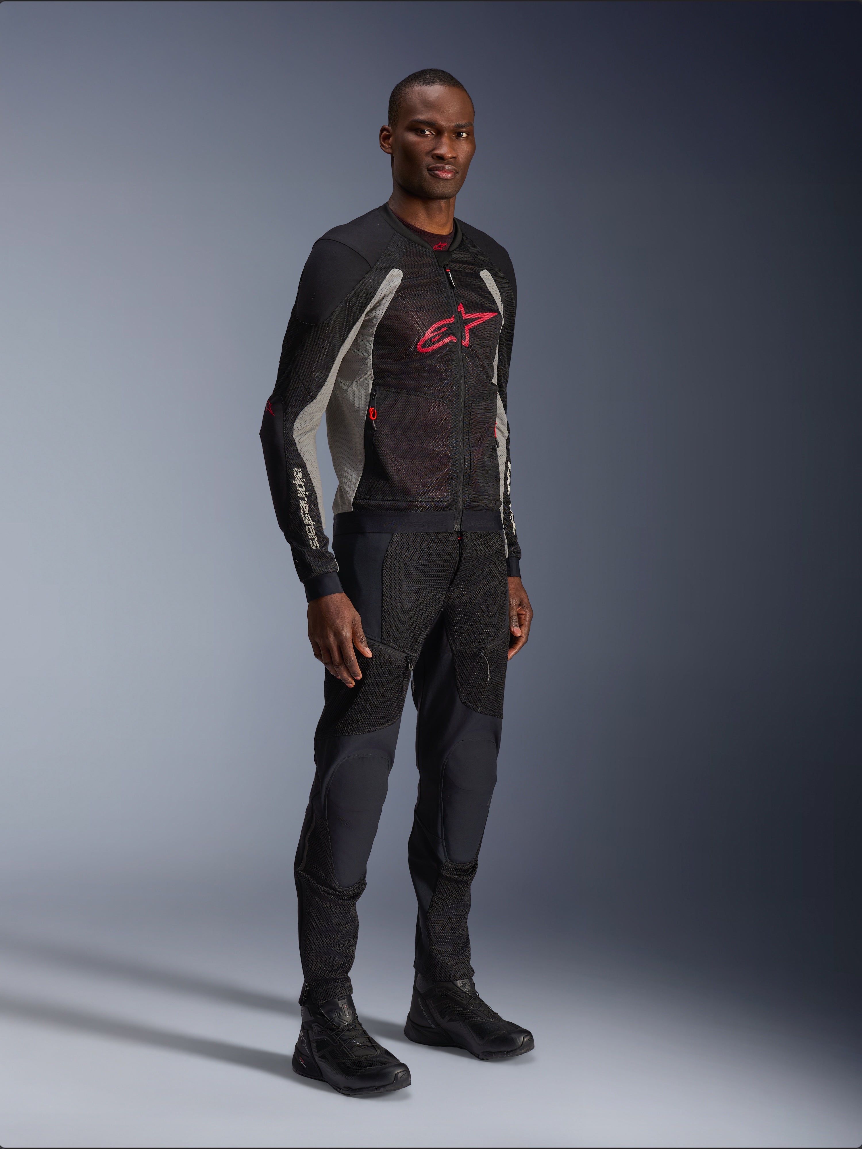 Alpinestars Troop-Air Jas, technische mesh motorjas in zwart, asgrijs en helderrood, gedragen door een model met bijpassende zwarte broek en laarzen, met een rood Alpinestars-logo op de borst, urban contemporary motorkleding