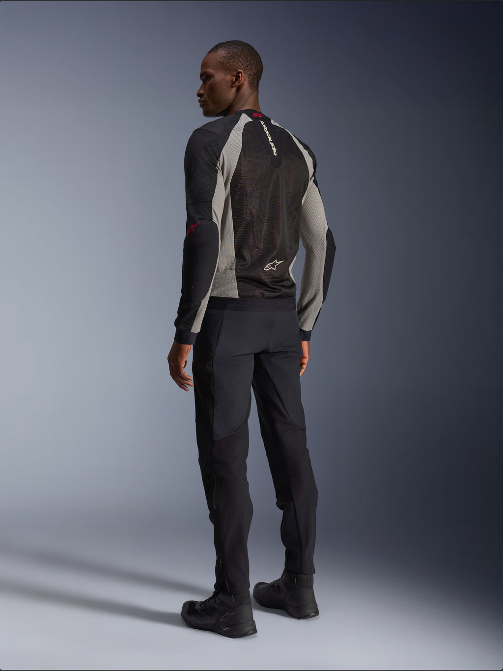 Model draagt de Alpinestars Troop-Air Jacket van achteren gezien, Black Ash Gray met felrode accenten, technische mesh-constructie voor ventilatie, gecombineerd met een zwarte motorbroek en motorlaarzen, urban contemporary stijl.