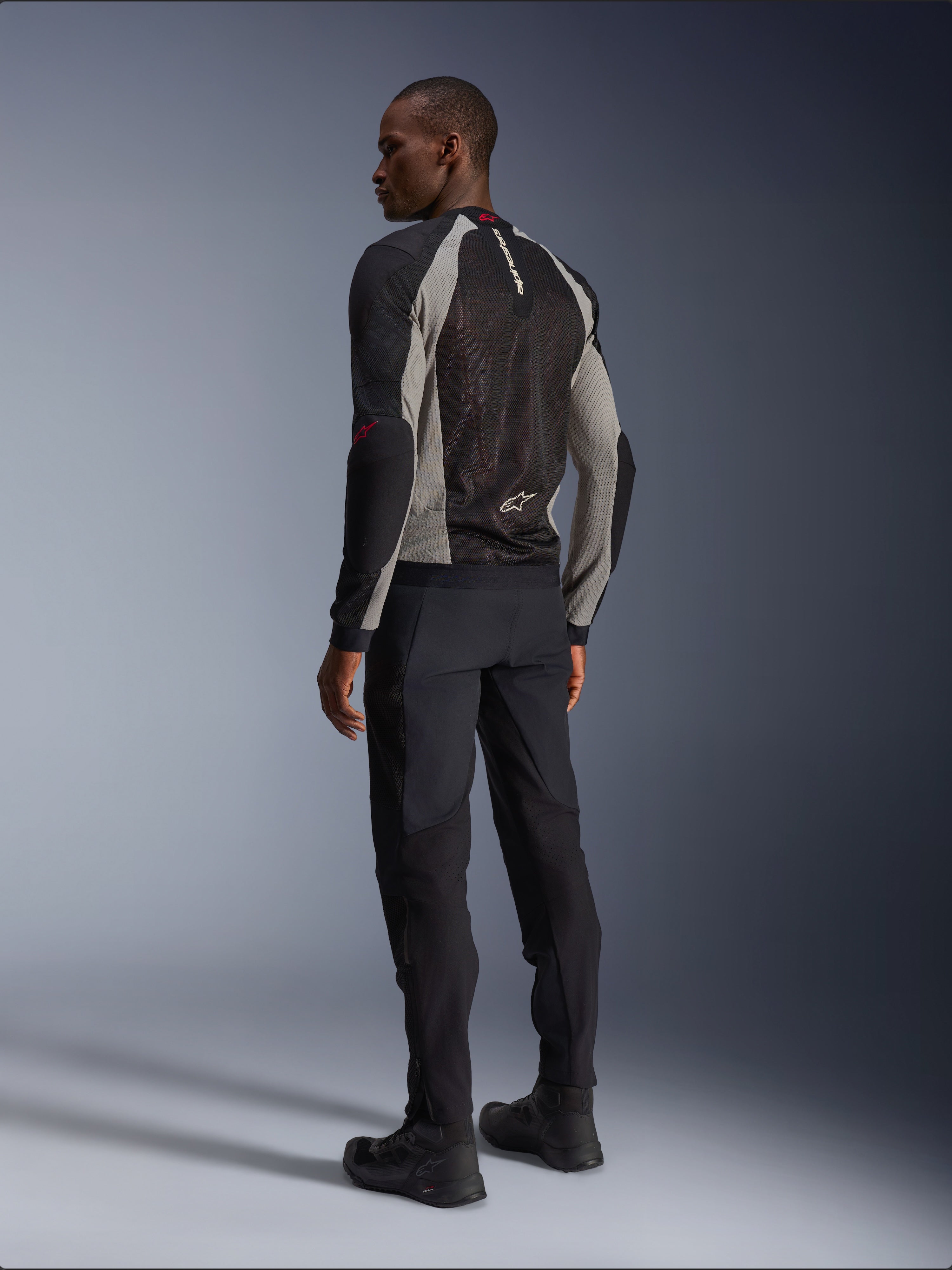 Model draagt de Alpinestars Troop-Air Jacket van achteren gezien, Black Ash Gray met felrode accenten, technische mesh-constructie voor ventilatie, gecombineerd met een zwarte motorbroek en motorlaarzen, urban contemporary stijl.