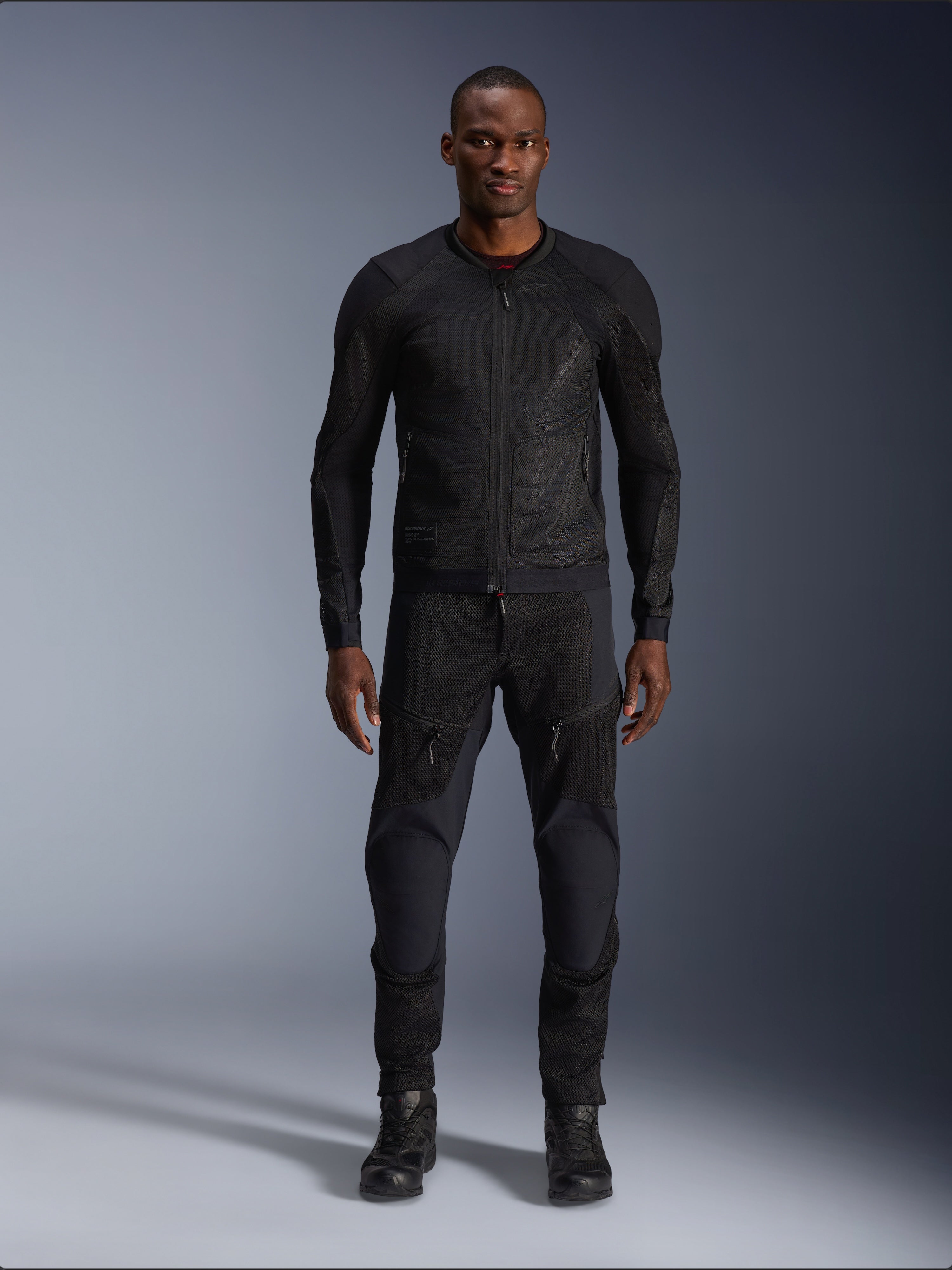 Mannelijk model in de Alpinestars Troop-Air Jas in Black Black, een lichtgewicht mesh motorjas gecombineerd met een bijpassende zwarte motorbroek en laarzen, voorzien van een technische 2D tech mesh-constructie voor superieure luchtstroom.