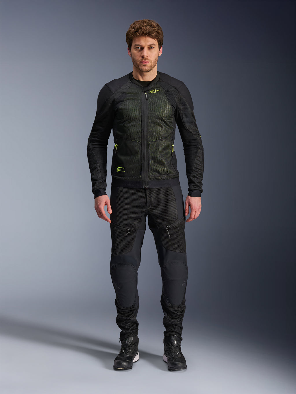 Mannelijk model draagt Alpinestars Troop-Air Jacket in Black Yellow Fluo, voorzien van technische mesh-panelen en felgele accenten, gecombineerd met een zwarte motorbroek en schoenen, eigentijdse motorkleding voor warm weer