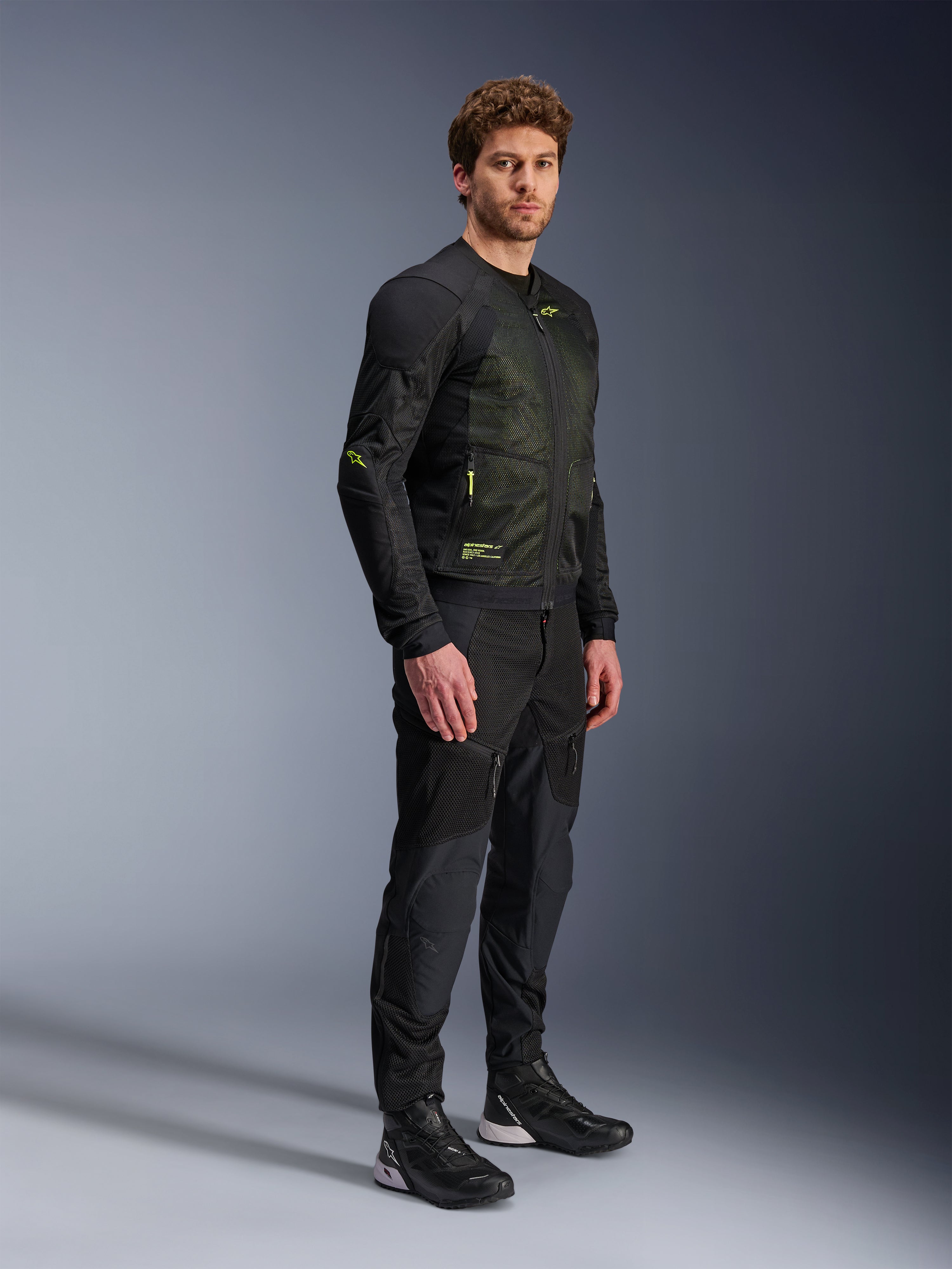 Alpinestars Troop-Air jas, mesh motorjas, zwart en fluogeel, gedragen door een model met bijpassende zwarte broek en motorschoenen, voorzien van mesh-constructie met hoge luchtstroom en fluorescerende gele accenten op de borst en mouwen, lichtgewicht technisch ontwerp voor rijden op de weg
