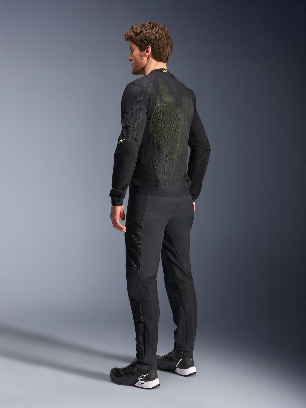 Alpinestars Troop-Air Jas in Black Yellow Fluo, achteraanzicht van een mannelijk model dat de technische luchtjas draagt, met een groot mesh rugpaneel met gele accenten en fluogele Alpinestars-logo's, gecombineerd met een zwarte motorbroek.