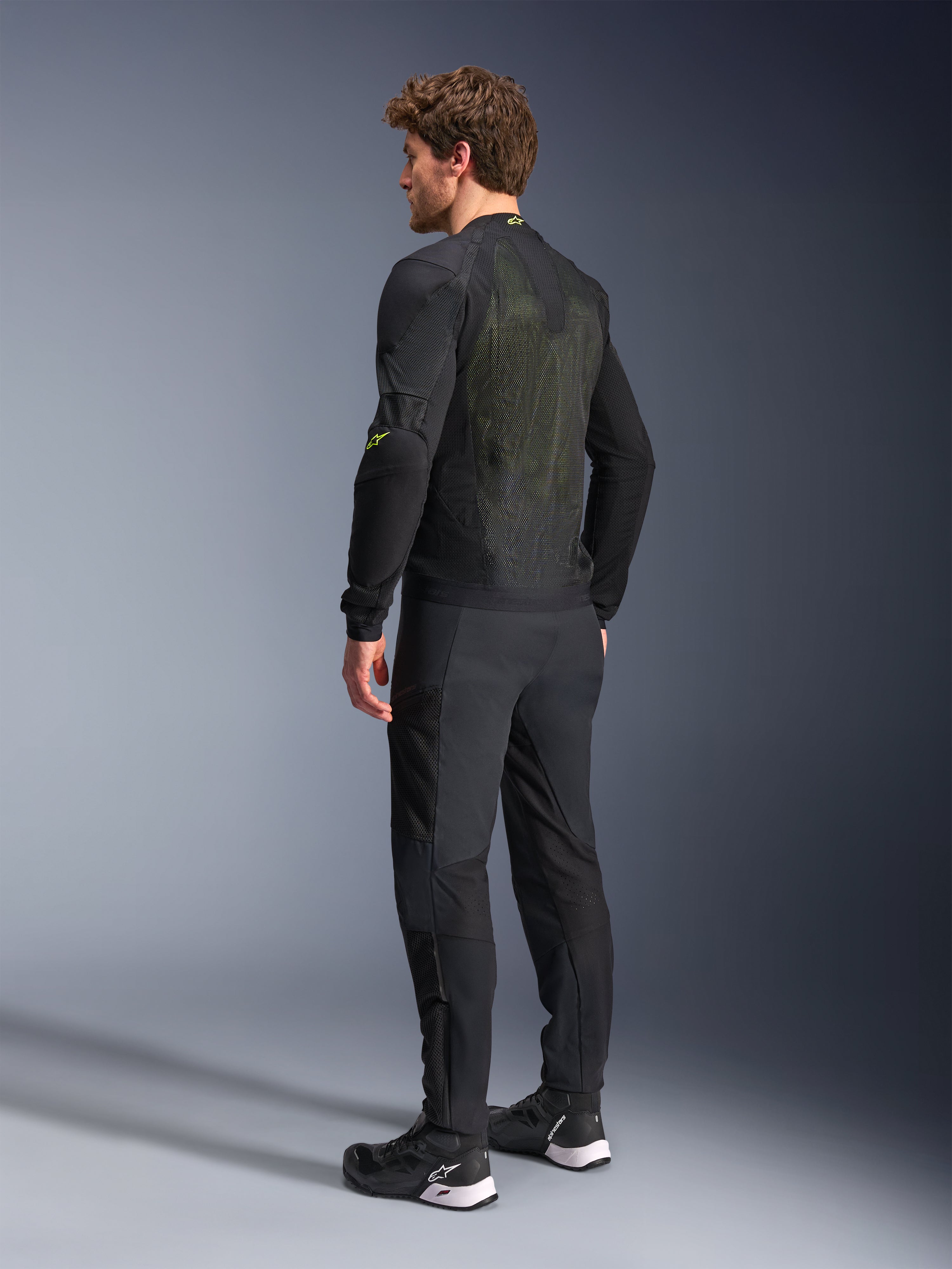 Alpinestars Troop-Air Jas in Black Yellow Fluo, achteraanzicht van een mannelijk model dat de technische luchtjas draagt, met een groot mesh rugpaneel met gele accenten en fluogele Alpinestars-logo's, gecombineerd met een zwarte motorbroek.