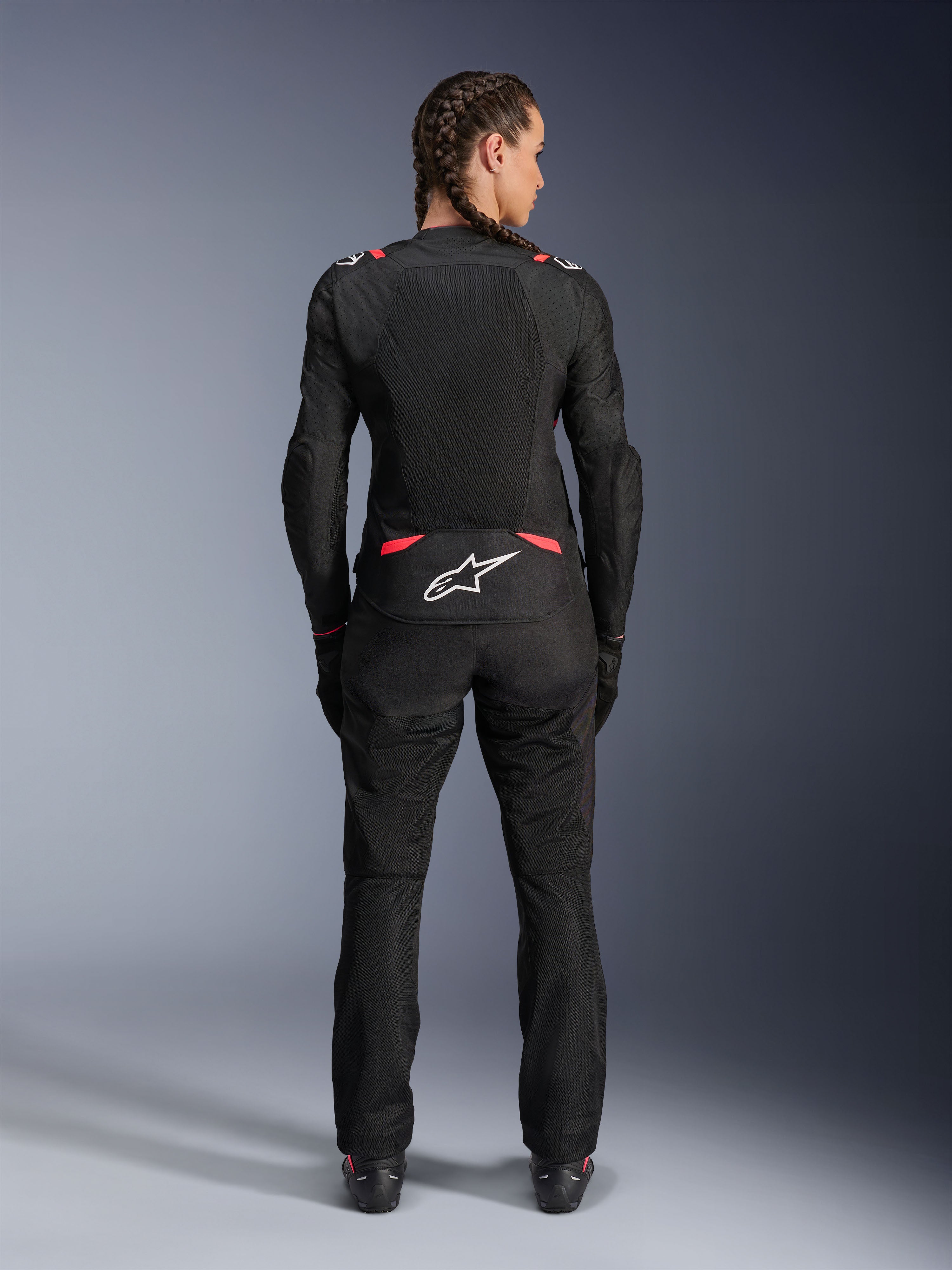Alpinestars Stella T-SPS Air V2 Jas, textiele motorjas voor dames, zwart en Diva Pink, achteraanzicht op model met ingevlochten haar, voorzien van grote mesh-panelen voor ventilatie, wit Astars-logo op de onderrug, gecombineerd met bijpassende zwarte motorbroek.