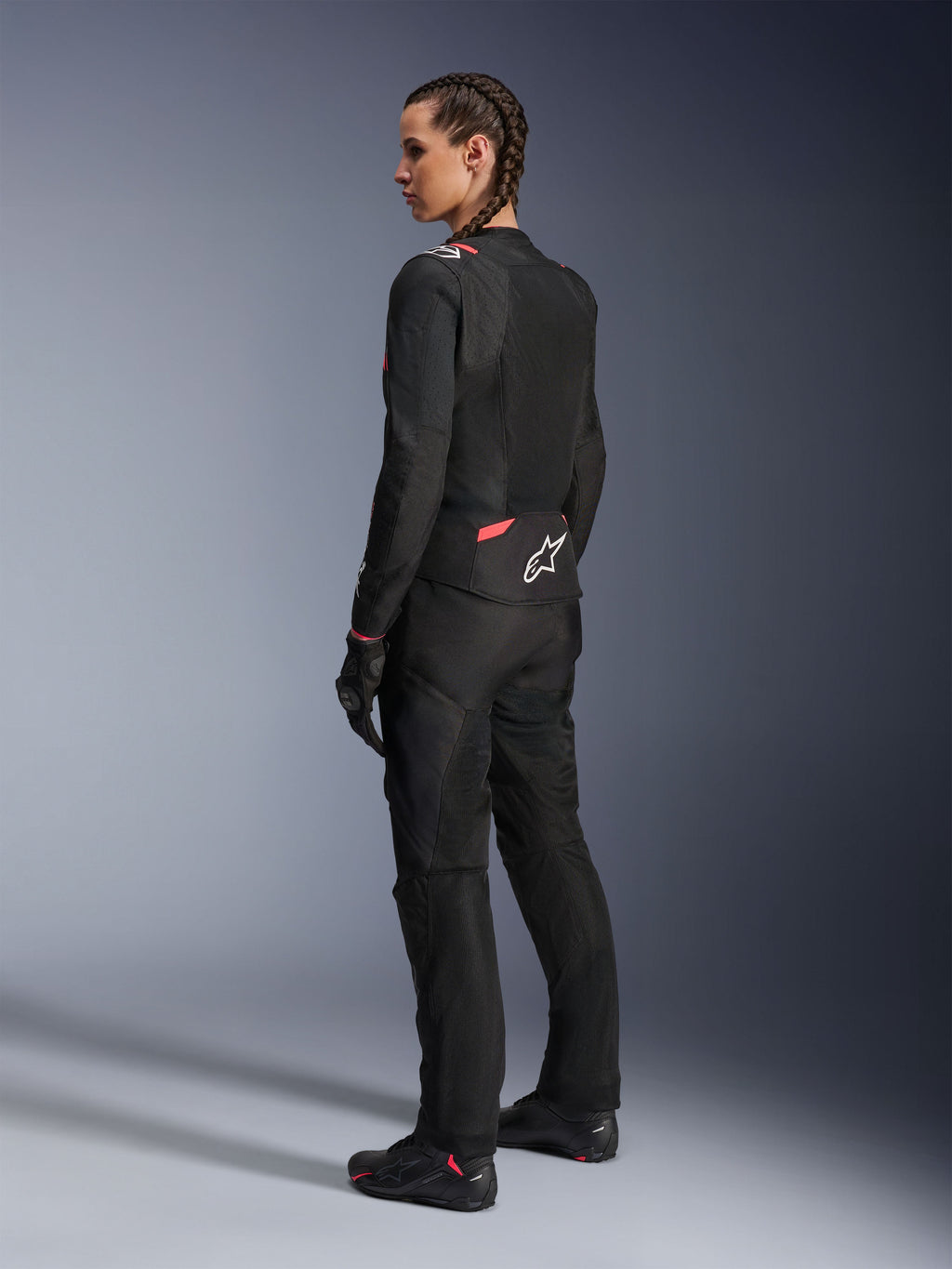 Alpinestars Stella T-Sps Air v2 damesmotorjas, zwart en Diva Pink, textiele motorjas getoond vanuit een schuin achteraanzicht, voorzien van grote mesh-panelen voor ventilatie en een wit sterlogo op de rug, gedragen met bijpassende zwarte motorbroek, handschoenen en laarzen