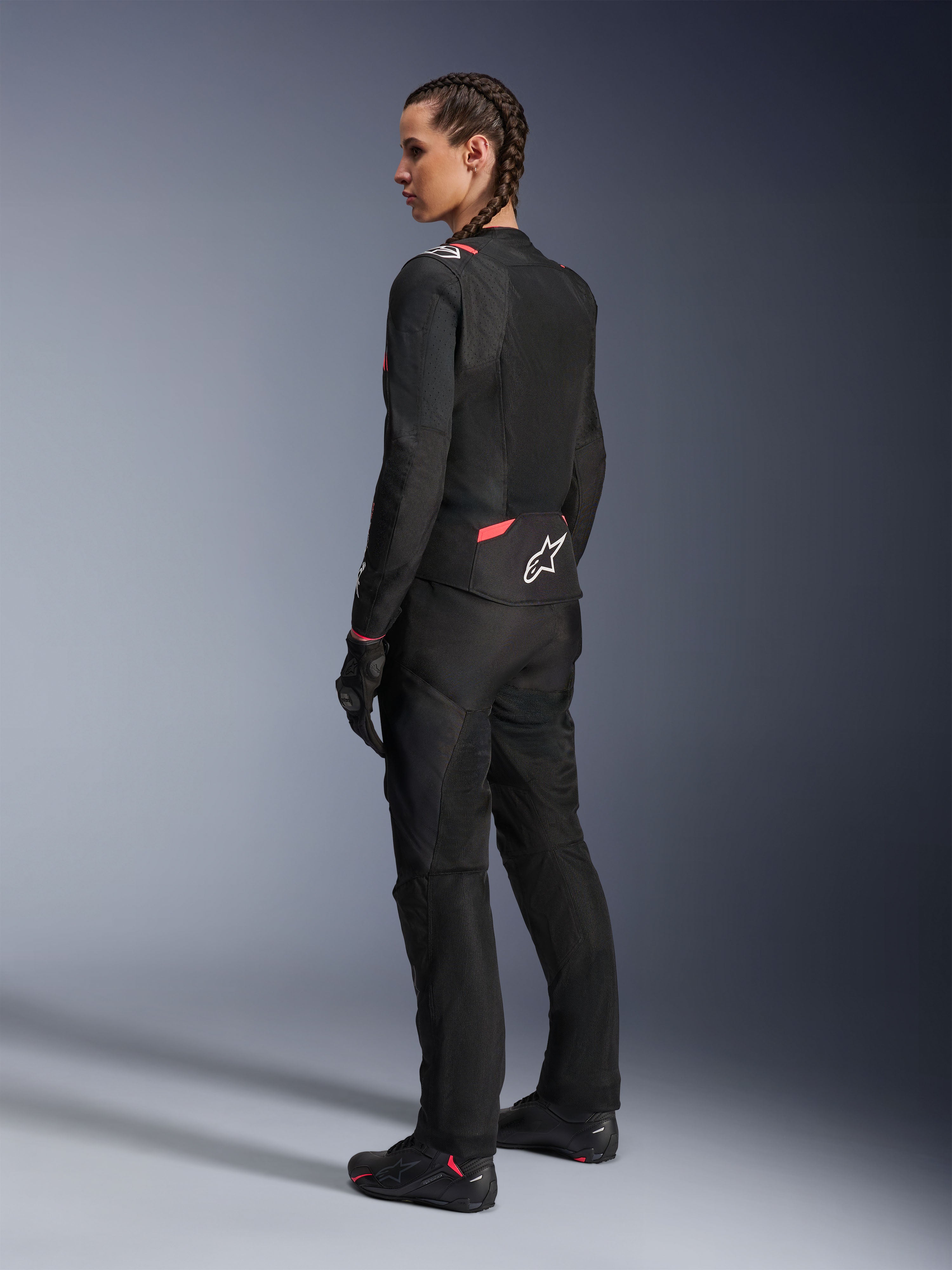 Alpinestars Stella T-Sps Air v2 damesmotorjas, zwart en Diva Pink, textiele motorjas getoond vanuit een schuin achteraanzicht, voorzien van grote mesh-panelen voor ventilatie en een wit sterlogo op de rug, gedragen met bijpassende zwarte motorbroek, handschoenen en laarzen