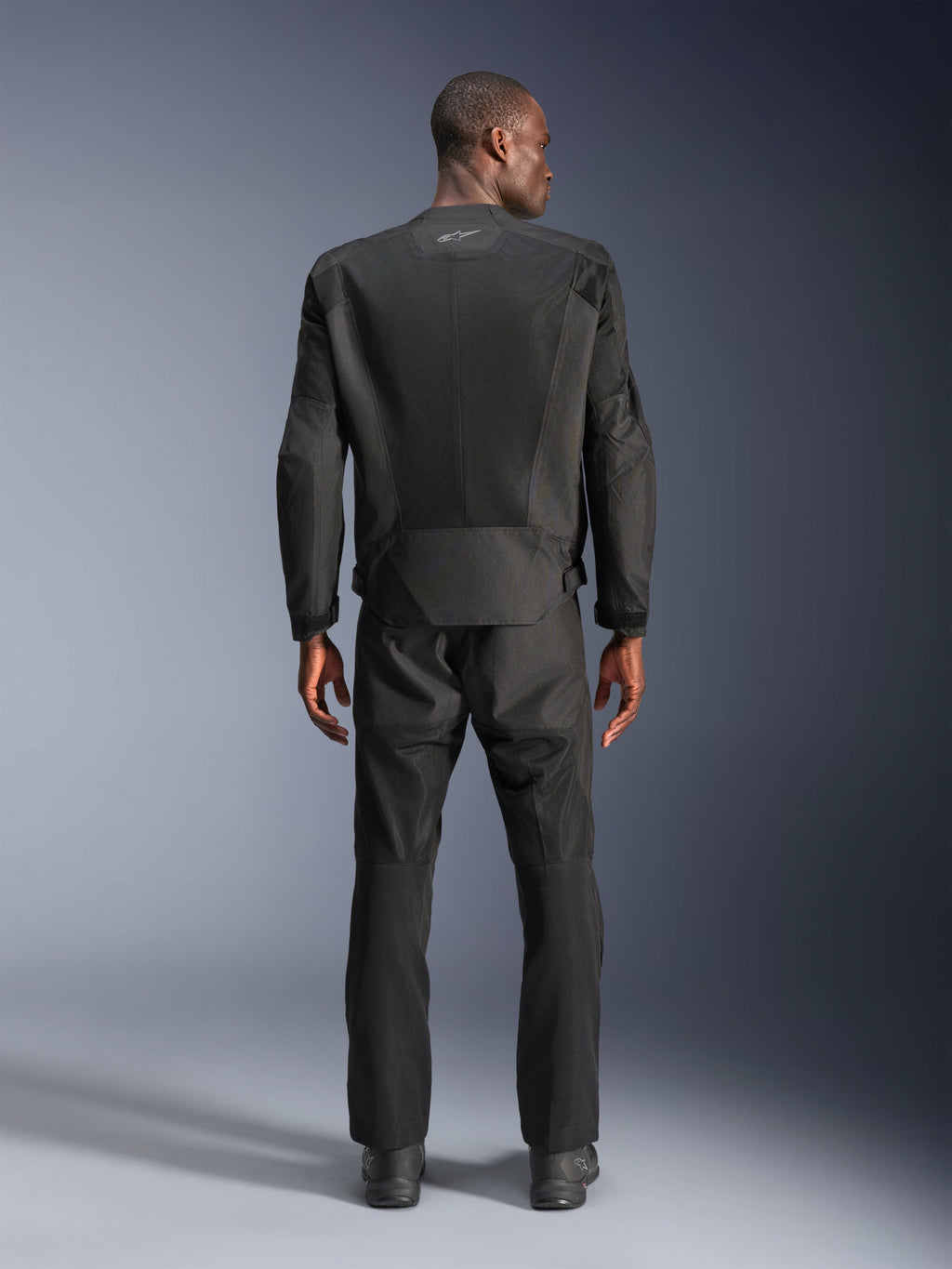Achteraanzicht van een man die de Alpinestars C-1 Air Jacket in het zwart draagt, urban motorjas met tech mesh panelen, bijpassende zwarte textiele broek en laarzen, strakke esthetiek met reflecterend logo op de bovenrug.