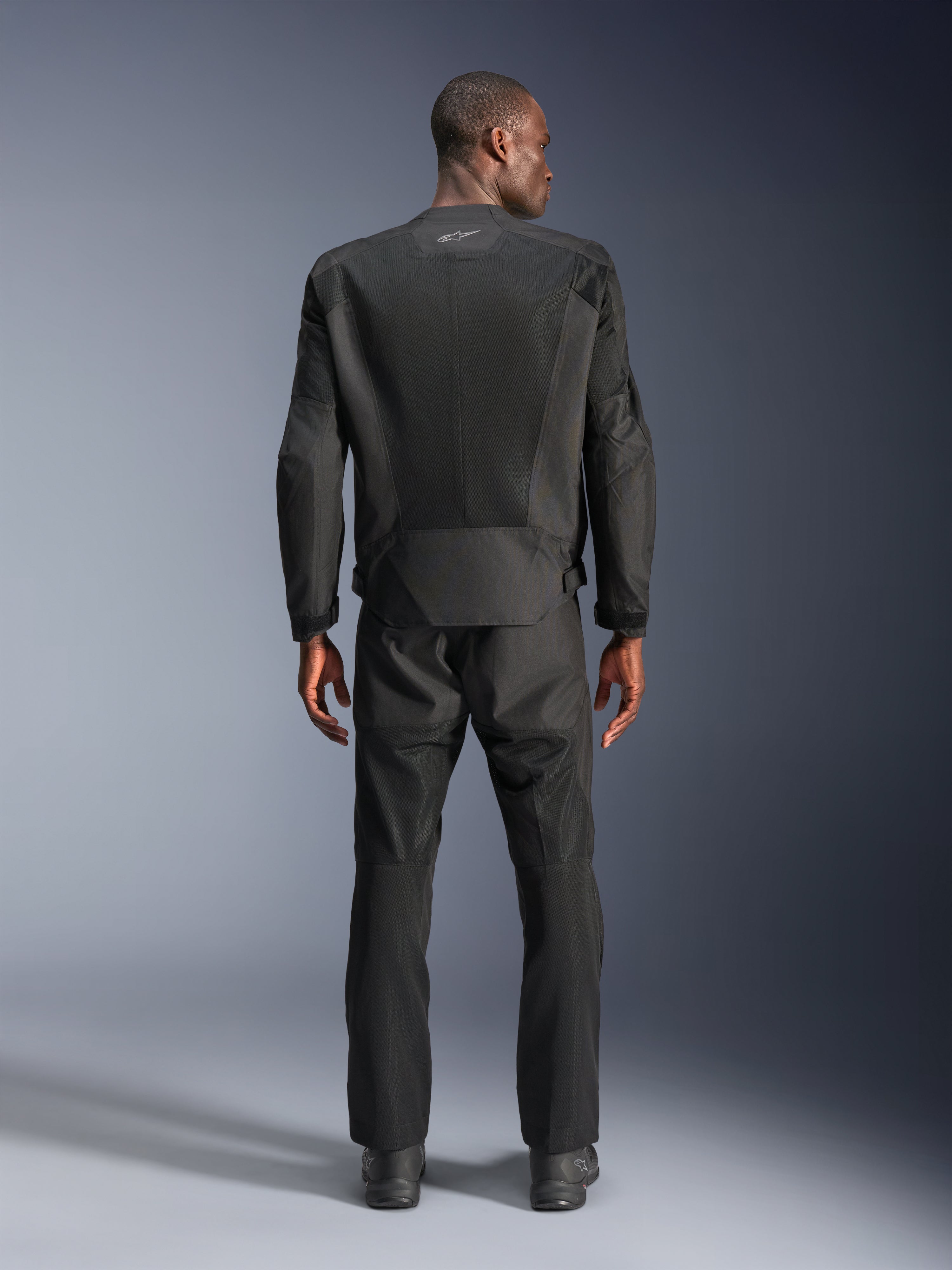Achteraanzicht van een man die de Alpinestars C-1 Air Jacket in het zwart draagt, urban motorjas met tech mesh panelen, bijpassende zwarte textiele broek en laarzen, strakke esthetiek met reflecterend logo op de bovenrug.