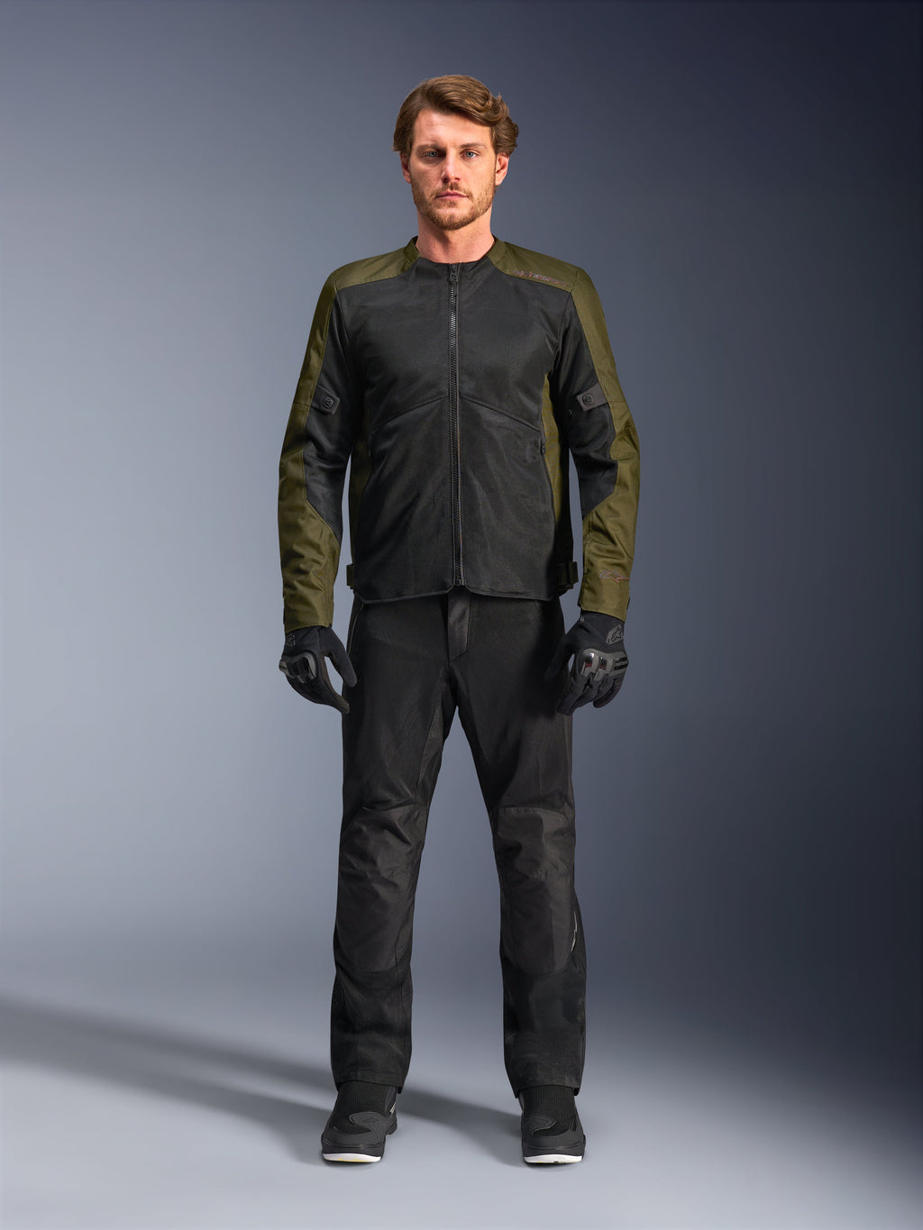 Alpinestars C-1 Air Jas, khaki en zwart, gedragen door een mannelijk model met een zwarte motorbroek en handschoenen, lichtgewicht textielontwerp met tech mesh-panelen, moderne stedelijke motoruitrusting